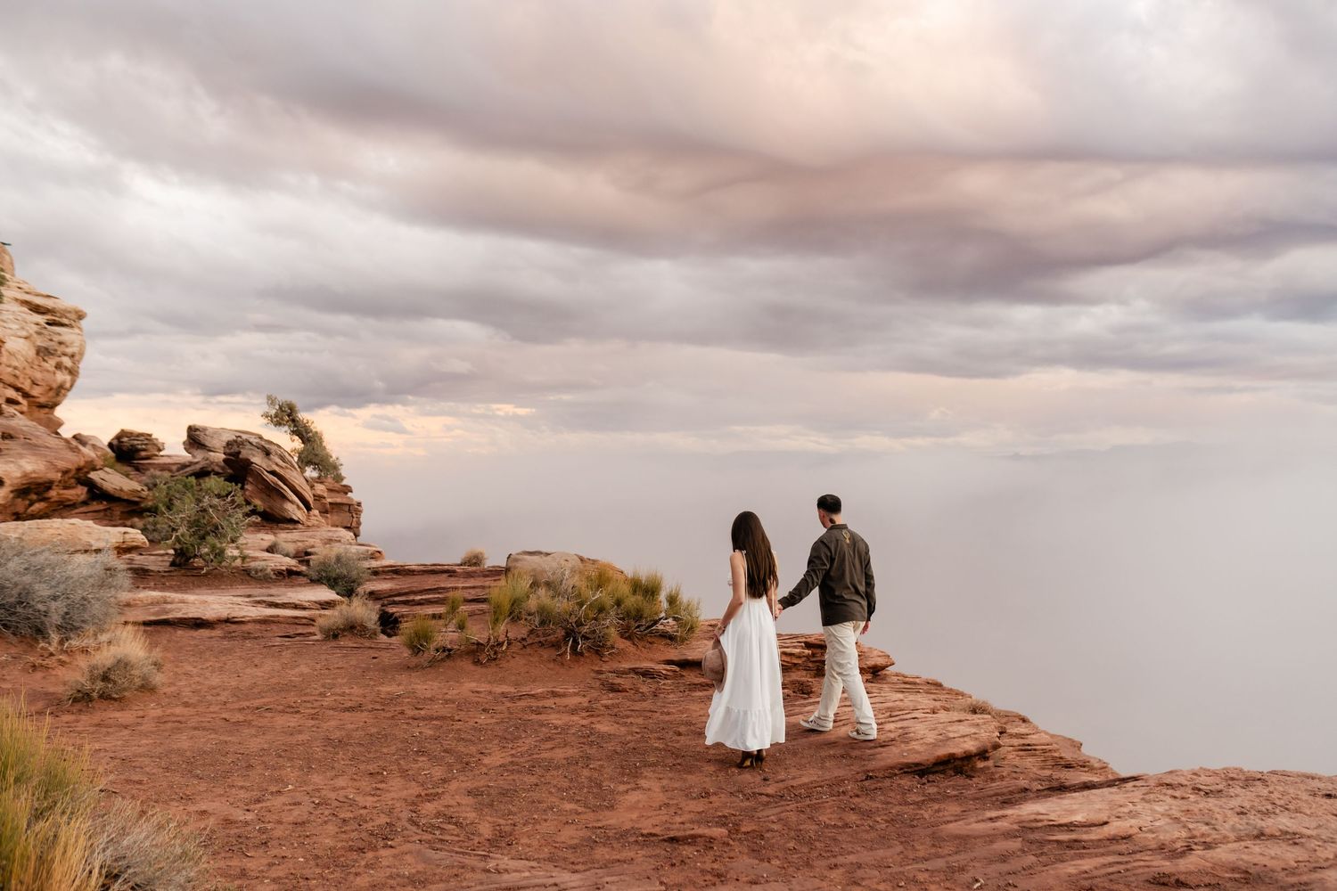 A couple walks along dramatic red rock cliffs at sunset overlooking a misty valley.
