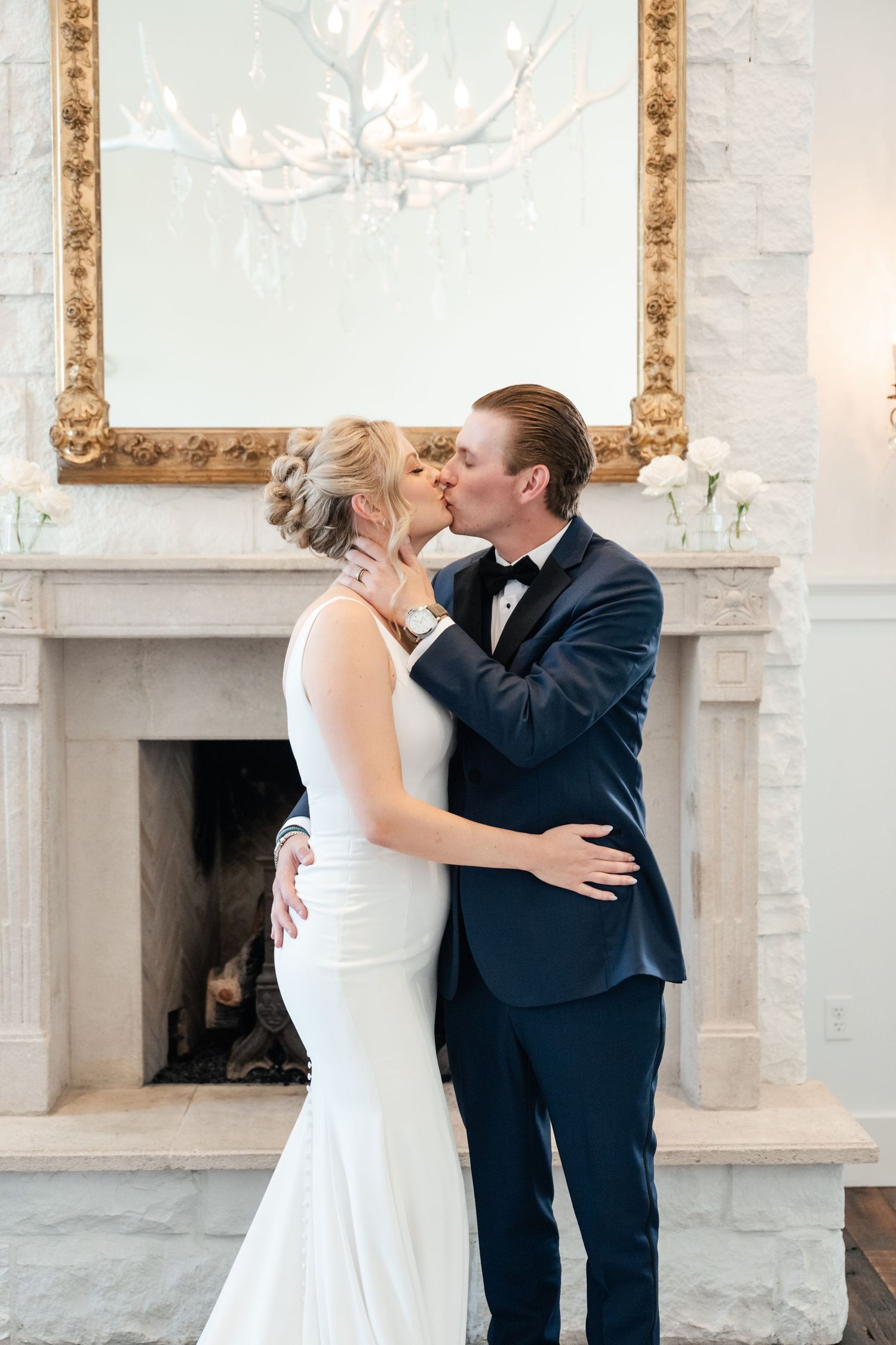 Bride and groom share a romantic kiss in front of an elegant fireplace in Park City, bride in white gown, groom in navy tuxedo.