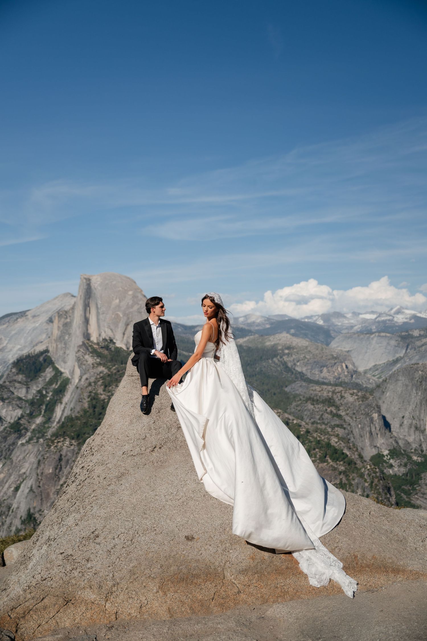 Bride and groom stand on rocky Glacier Point overlook in Yosemite, wedding dress flowing, Half Dome visible behind them.