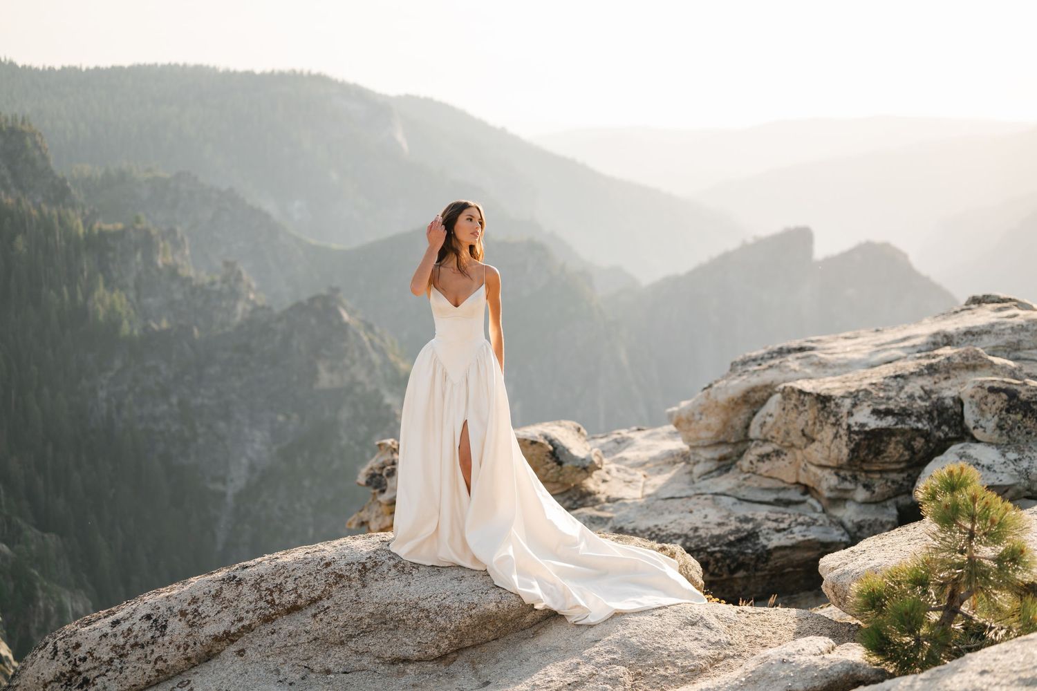 Woman in white wedding gown poses on rocky mountain overlook with stunning valley and peaks backdrop.