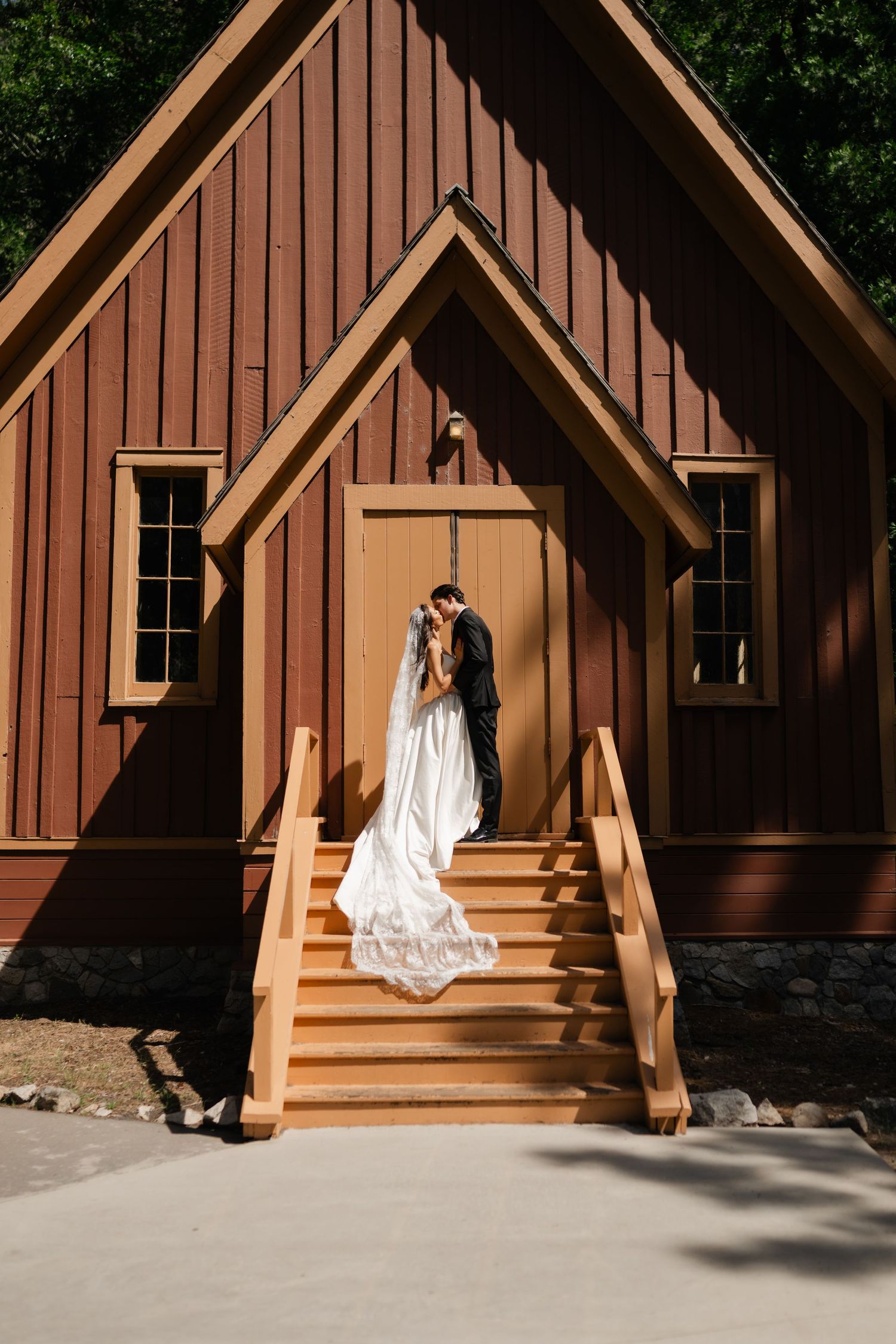 Bride and groom share a kiss on wooden chapel steps, bride's white gown trailing elegantly before a rustic A-frame building.