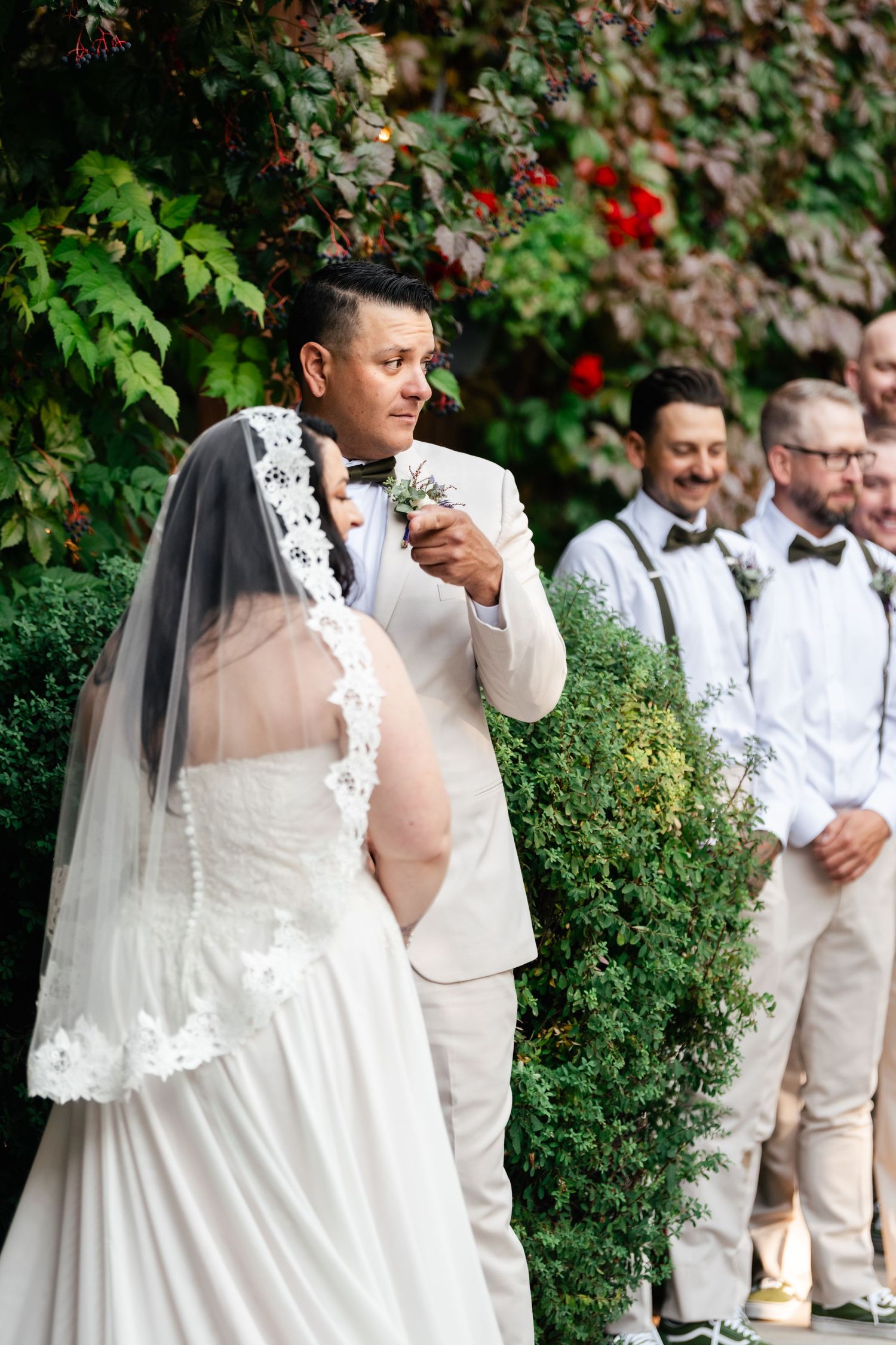A wedding couple stands in a garden setting surrounded by greenery and red flowers while groomsmen look on.