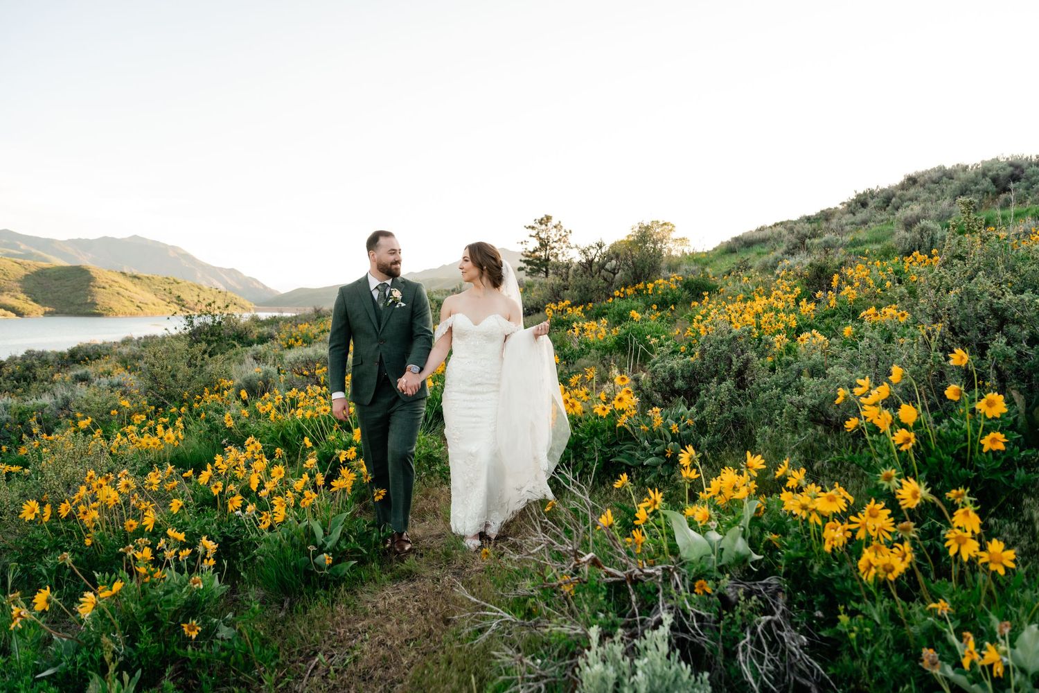 Couple walking along hillside trail through vibrant yellow wildflowers during spring in mountainous landscape.
