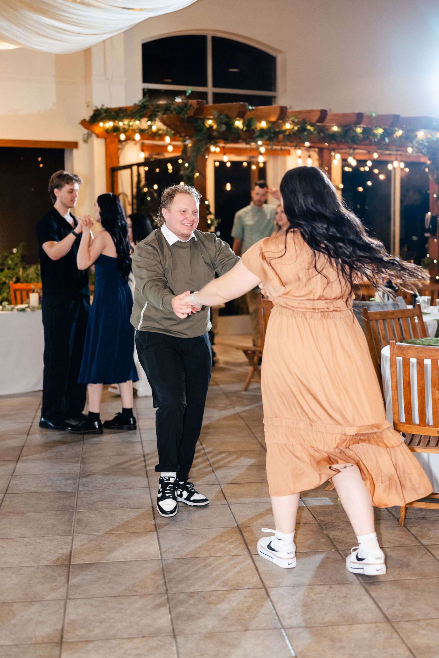 Two guests dance energetically on a tiled floor under string lights at a wedding reception.