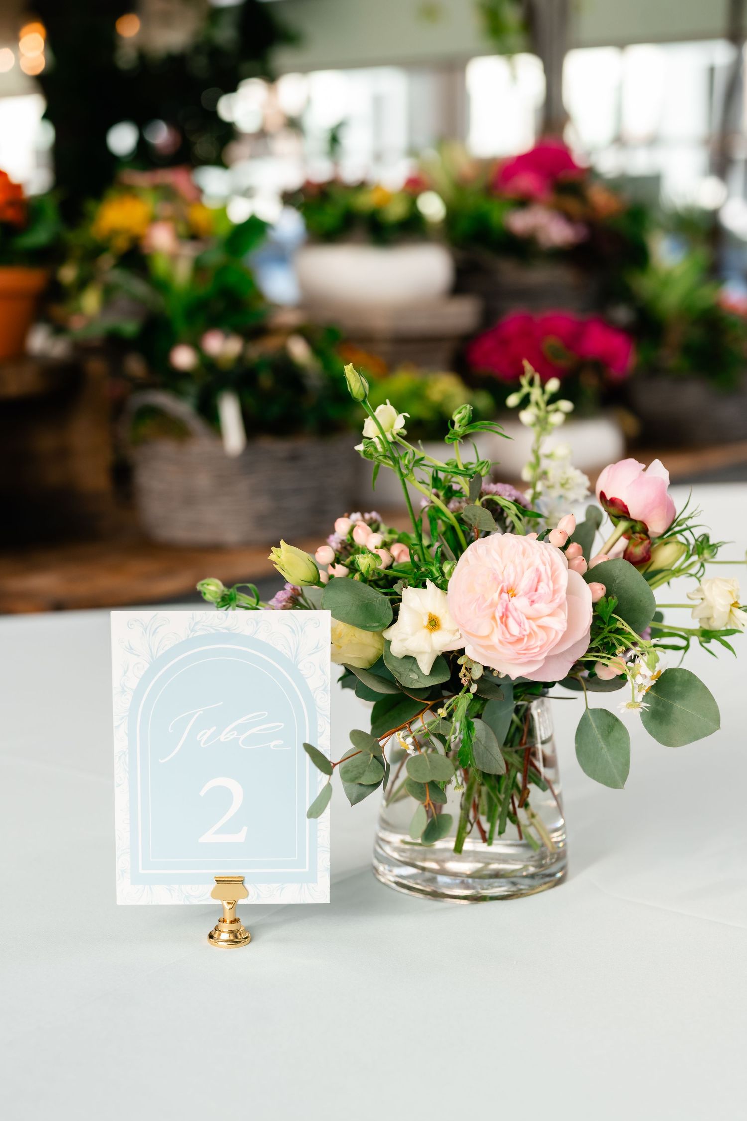 Elegant wedding table centerpiece with pink roses and eucalyptus in a glass vase next to a light blue table number card.