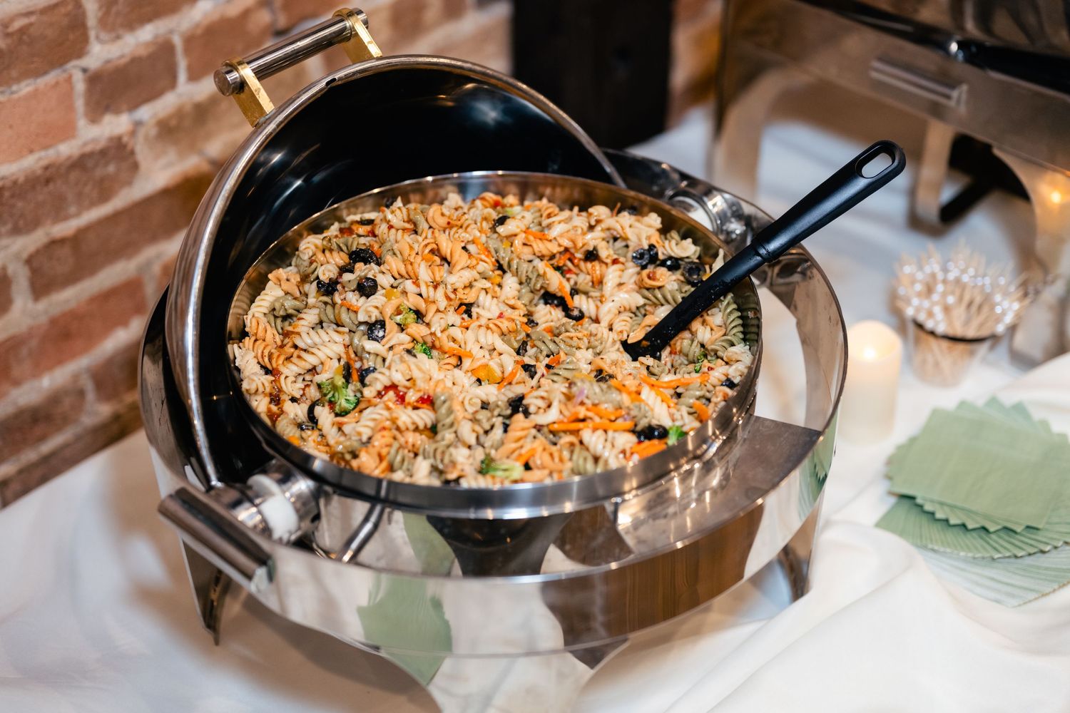 A large skillet filled with sauteed wild rice pilaf and vegetables sits on a buffet table with brick wall background.