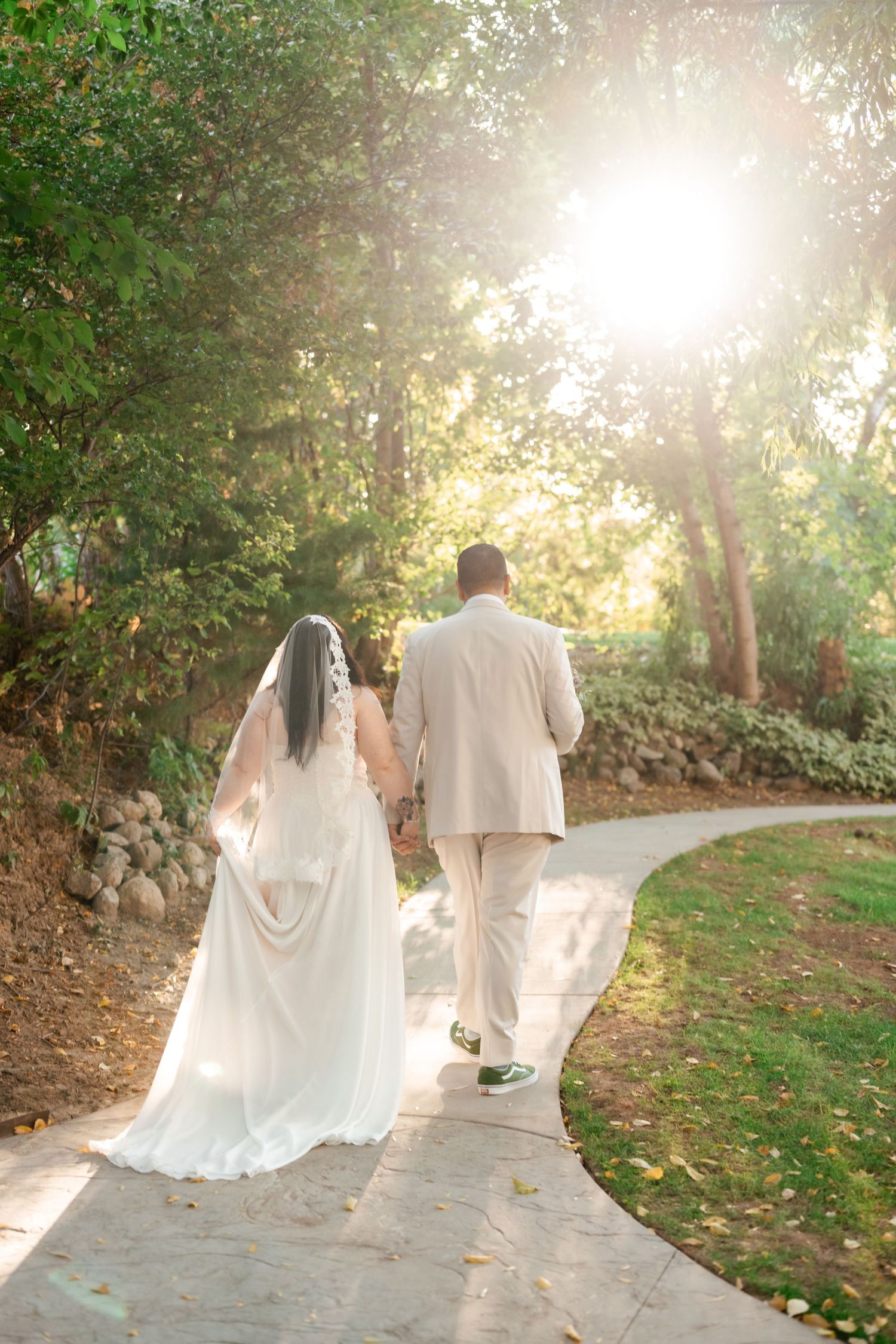 A romantic wedding couple walking down a sunlit garden path surrounded by lush greenery.
