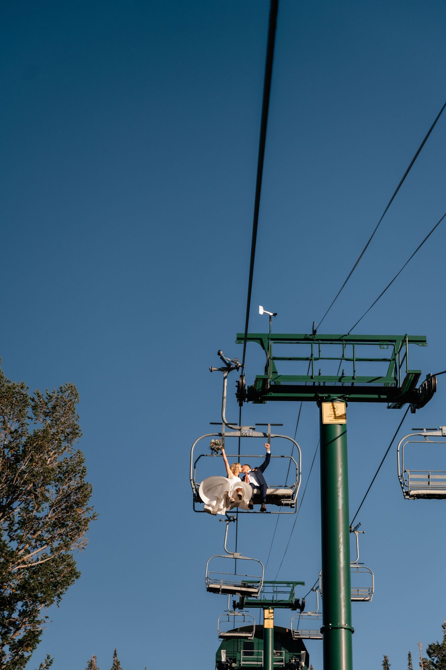 A wedding couple sit on a ski lift chair against a bright blue sky on a sunny day. Park City Wedding Photographer. Utah Wedding Photographer