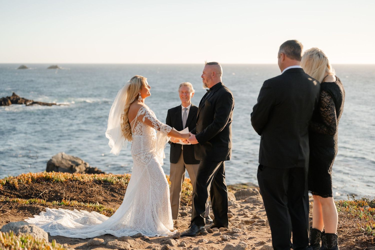 An intimate wedding ceremony takes place on a rocky coastal cliff overlooking the ocean at sunset.
