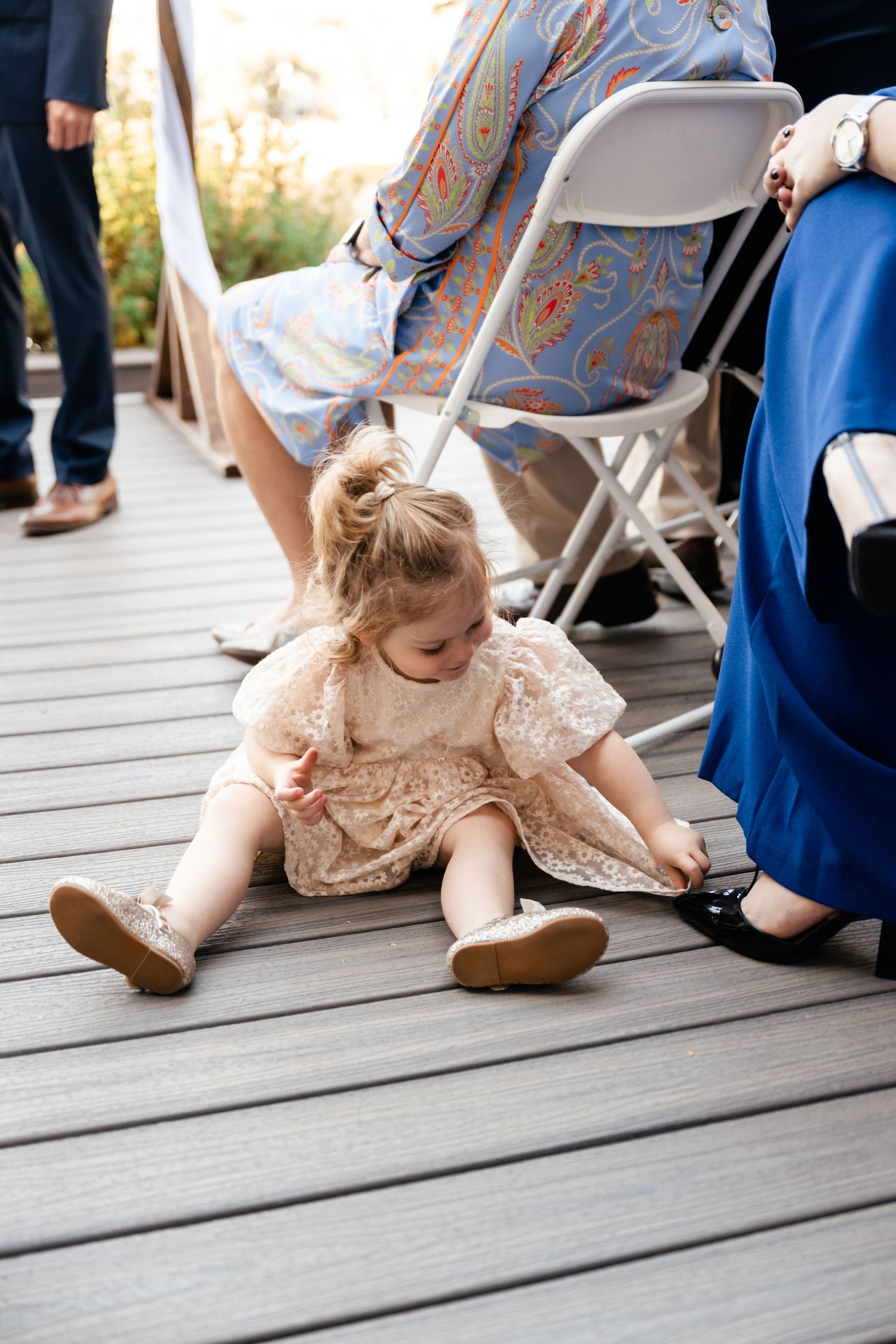 Small child in lace dress sitting on wooden deck during formal event.