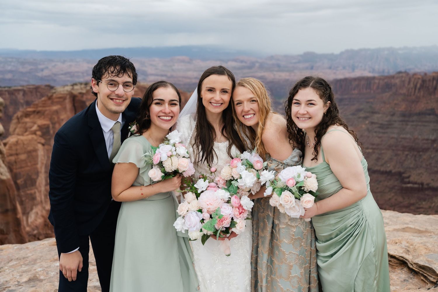 Wedding party in sage green and formal attire poses together against desert canyon backdrop.