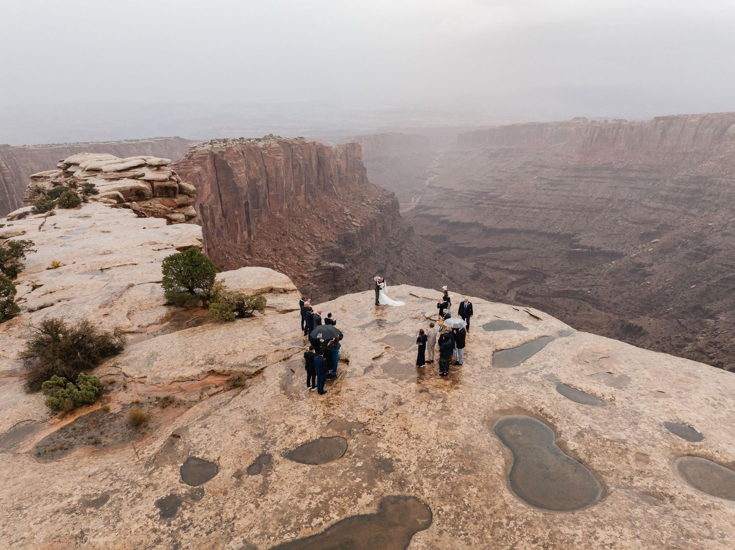 Visitors gather at scenic overlooks along the rim of a vast desert canyon on a misty overcast day.