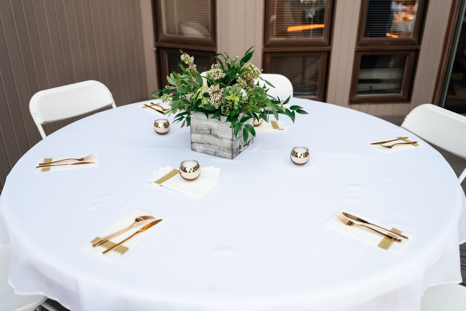 A round white table with place settings and a centerpiece of greenery in a wooden box.