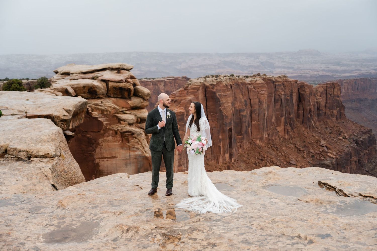 Wedding couple standing on the edge of the Grand Canyon with vast desert landscape and red cliffs in the background.