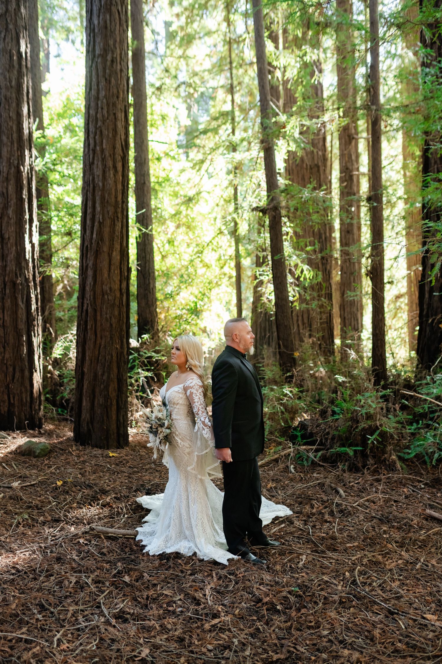 A couple in formal wedding attire standing among towering redwood trees.