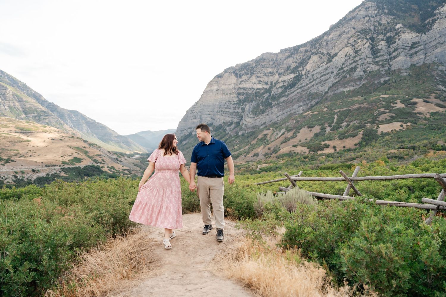 Two people walking hand in hand along a winding mountain path surrounded by rugged terrain and distant peaks.