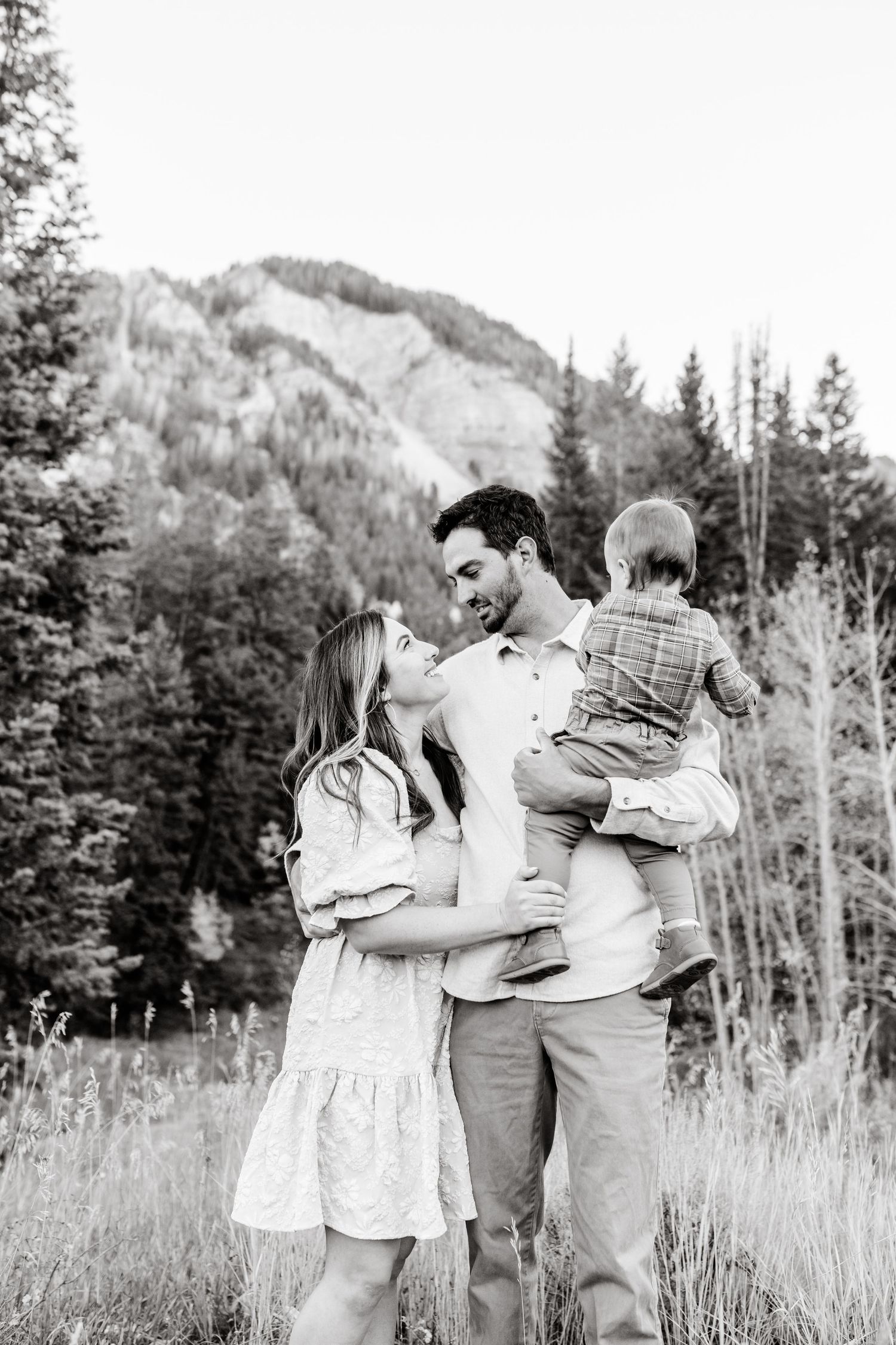 Family embracing in a scenic mountain meadow with evergreen trees and rolling hills in the background.