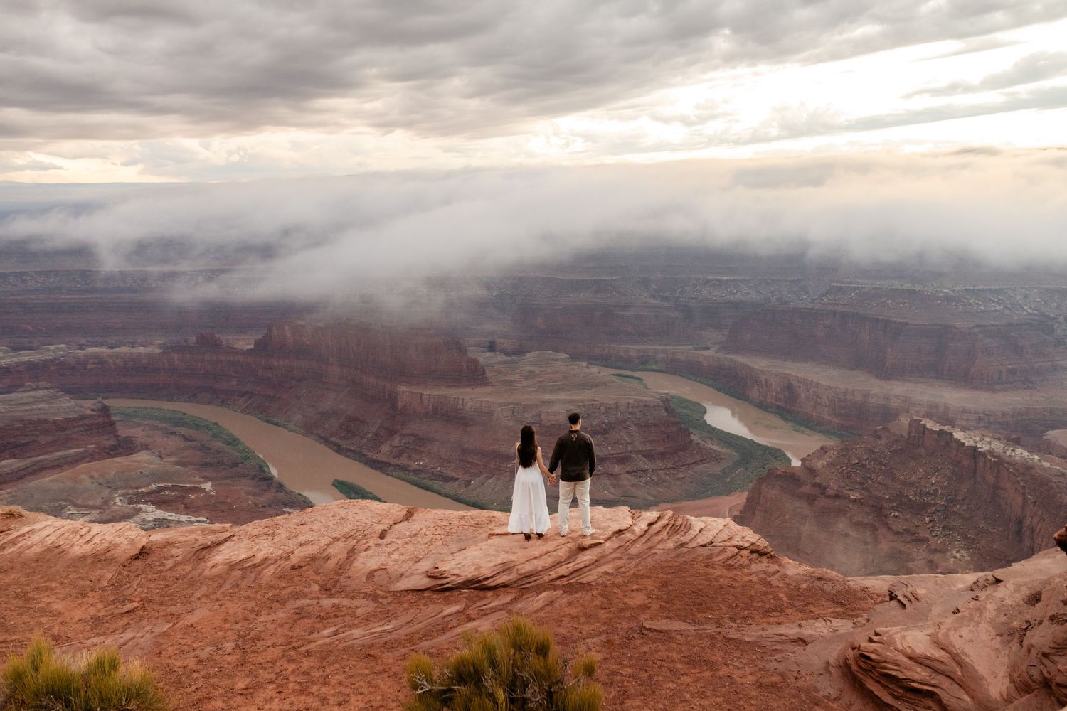 Panoramic view of a vast desert canyon landscape with dramatic cloudy skies and distant figures on the rim.