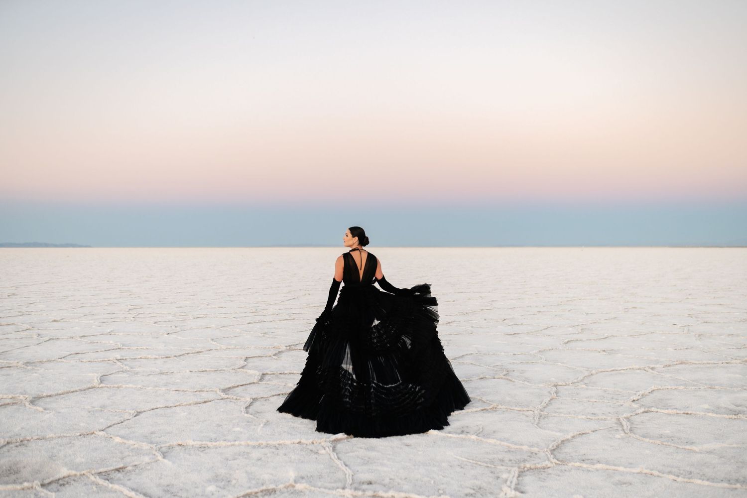 A figure in a flowing black dress walks across the stark white landscape of a salt flat at sunset. Utah Salt Flats Photographer.