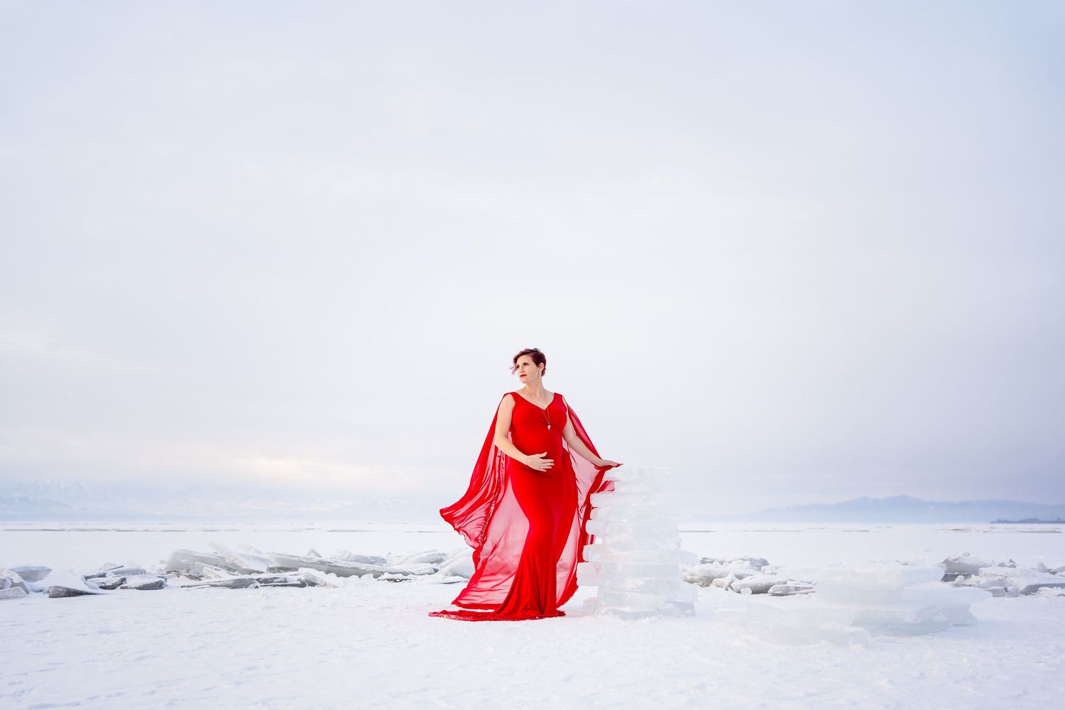 A person in a flowing red dress poses dramatically against a stark white snowy landscape at sunset.