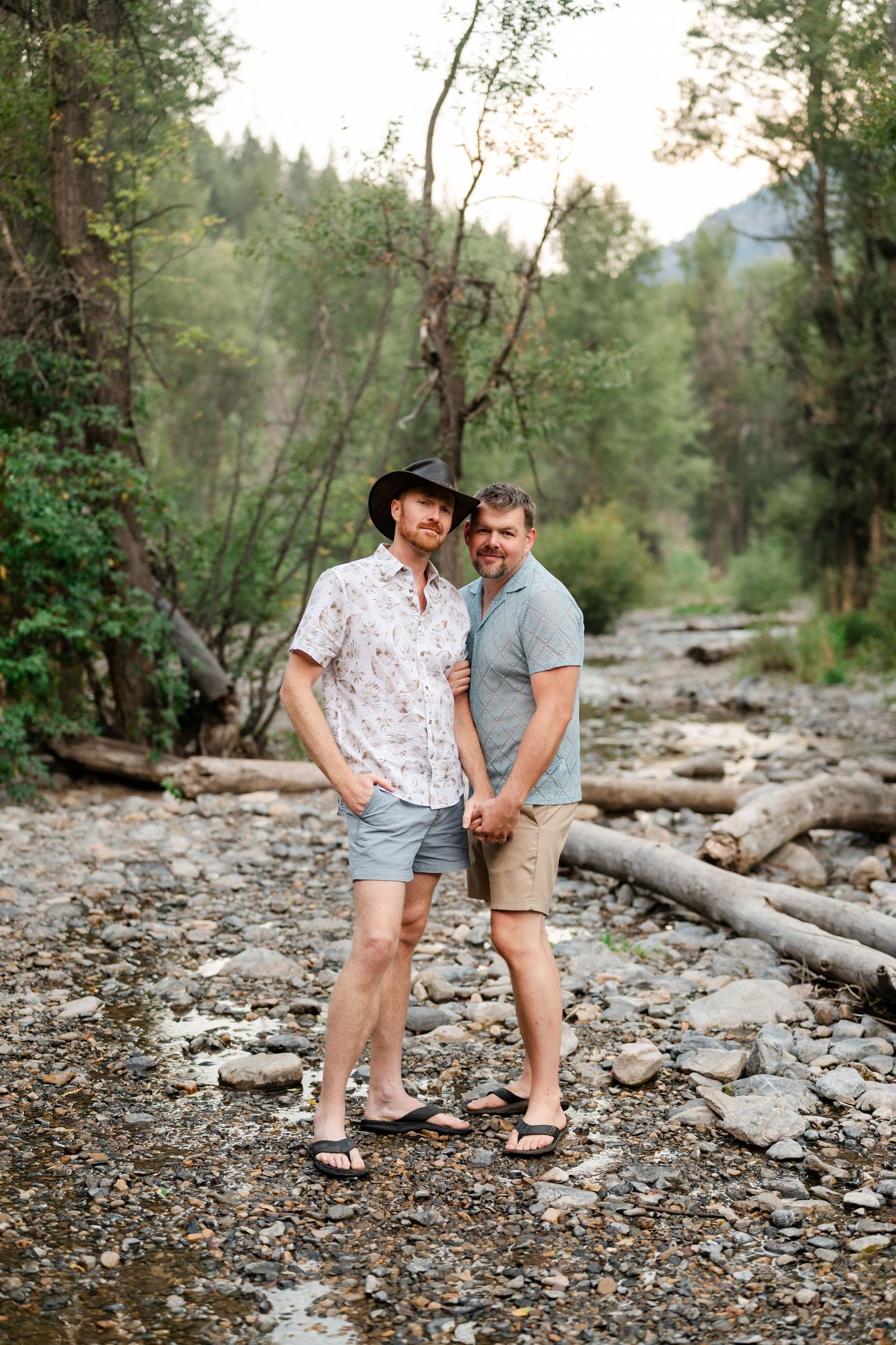 LGBTQ friendly engagement photographer, Two men standing together on a rocky riverbed surrounded by lush green trees and mountains.