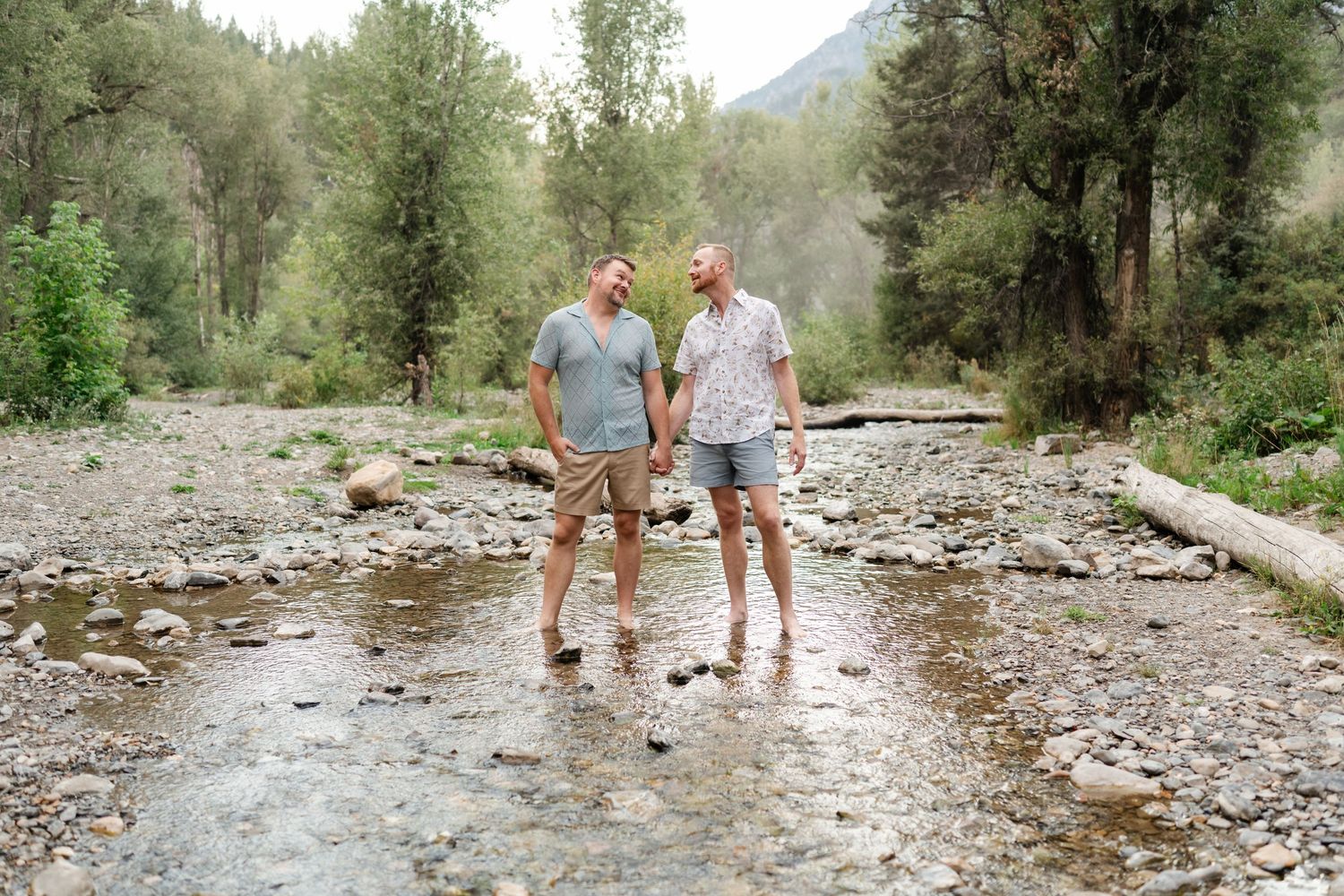 Gay couple holding hands and smiling at each other while standing in a rocky mountain stream surrounded by trees.