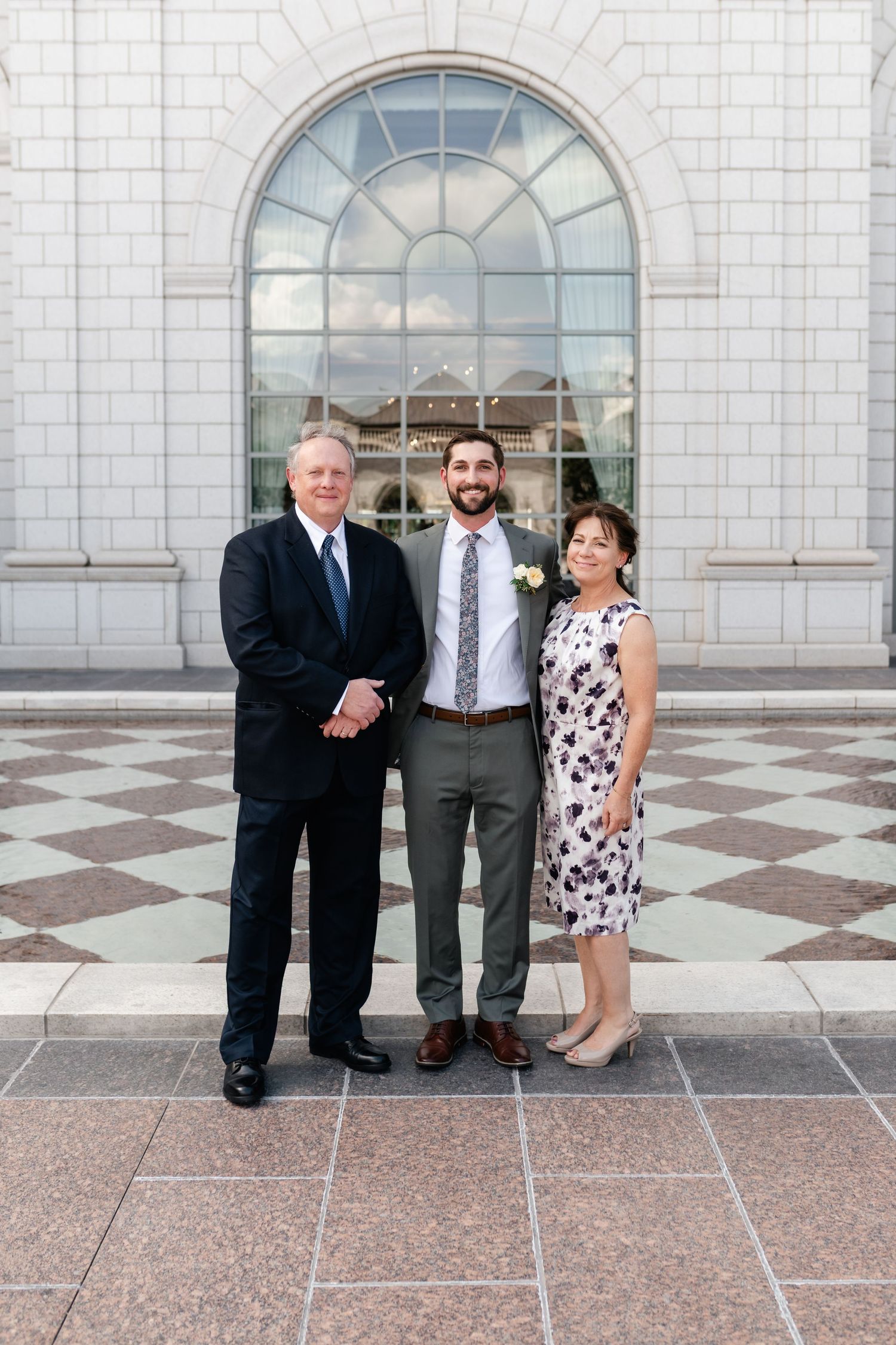 Three people pose together in formal attire against a white stone building with arched windows.