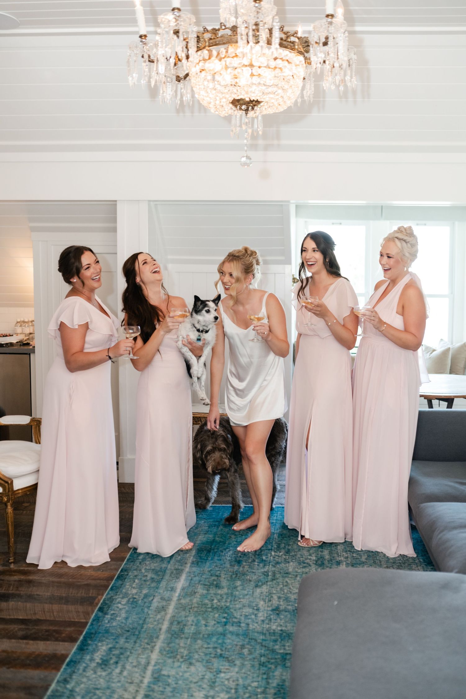 Group in blush pink bridesmaid dresses sharing a joyful moment with a dog on a blue rug.