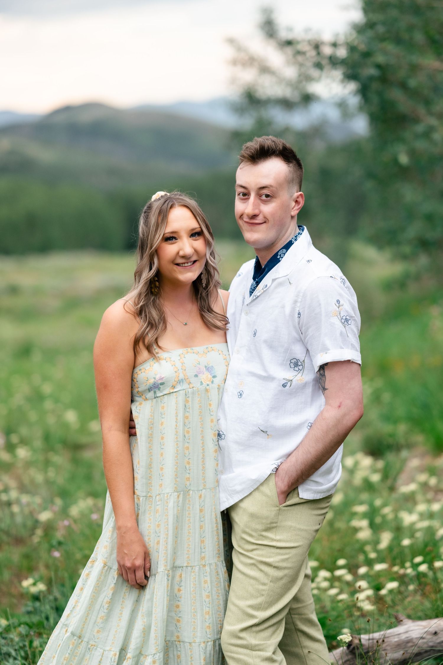 A couple poses together in a mountain meadow wearing light summer attire.