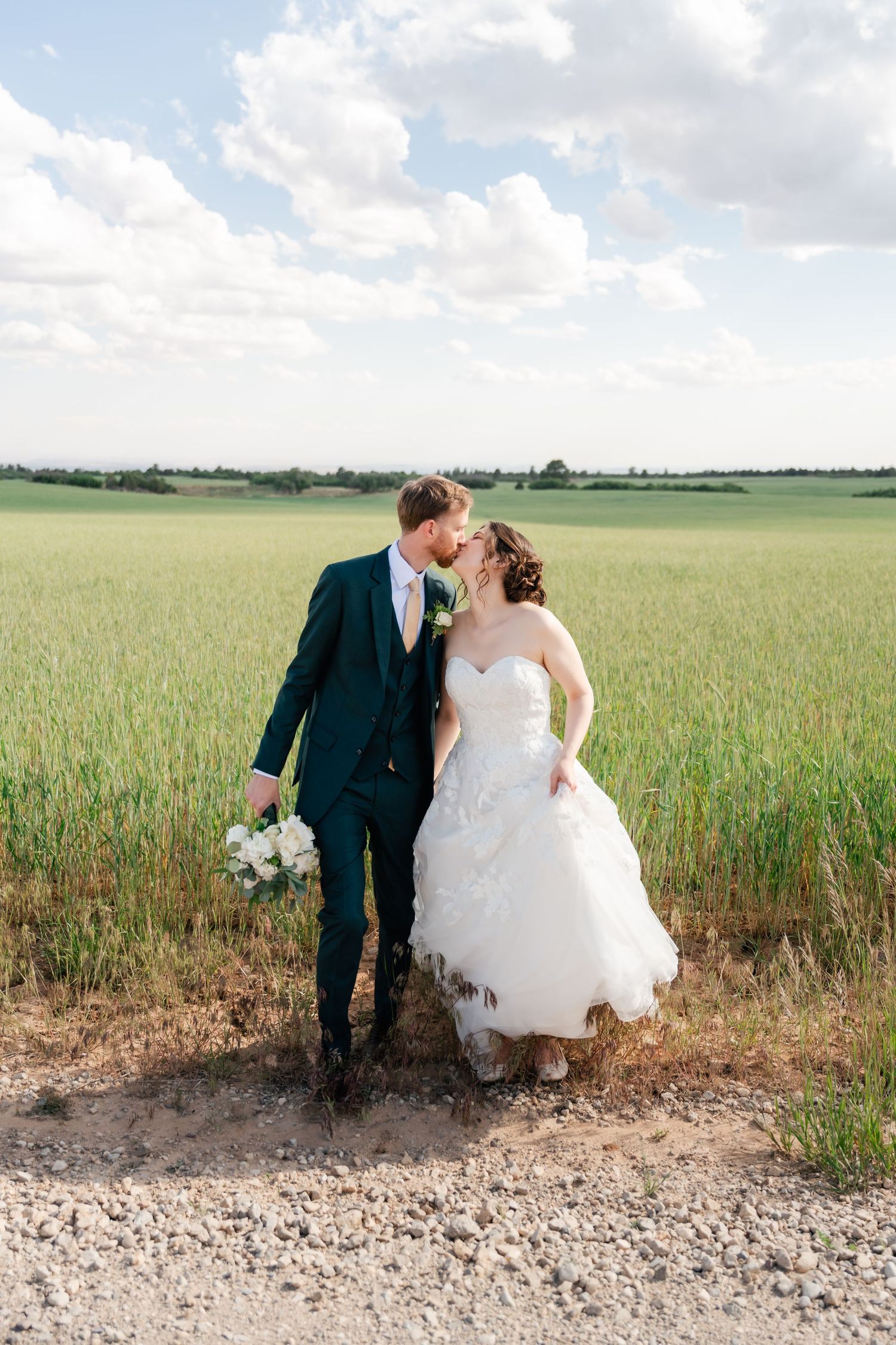 A couple poses in a scenic wheat field wearing formal wedding attire on a bright summer day.