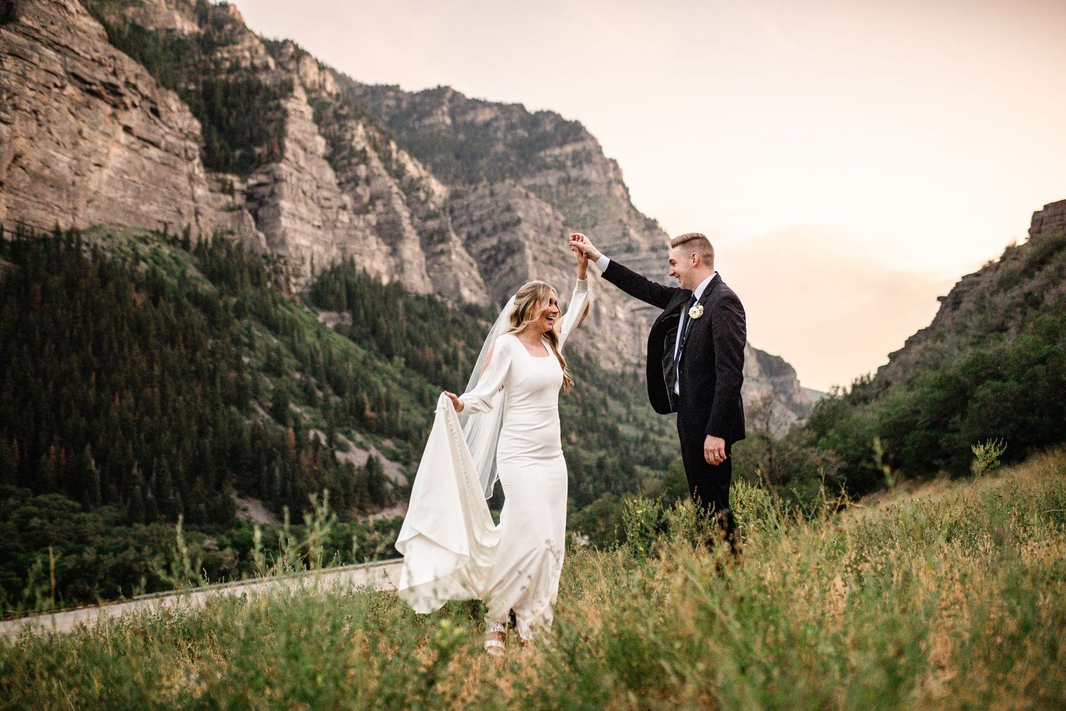 A couple in wedding attire stands together in a scenic mountain meadow during sunset. Utah Mountain Wedding Photographer.