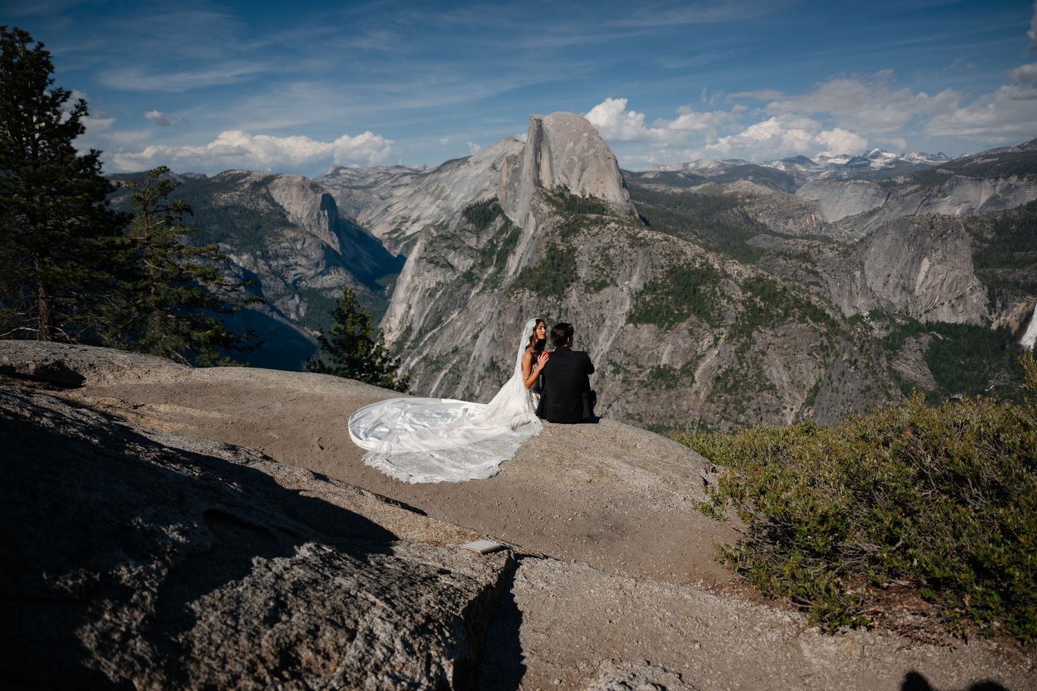 Bride and groom embrace on rocky overlook with Half Dome and Yosemite Valley in background on wedding day.