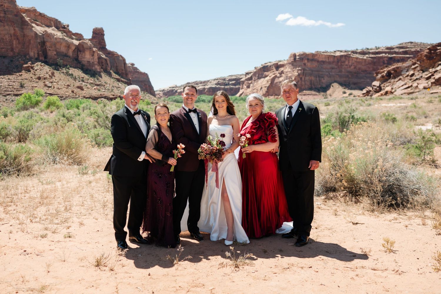 Wedding party posing in red rock desert landscape, bride in white gown with guests in burgundy and red formal attire.