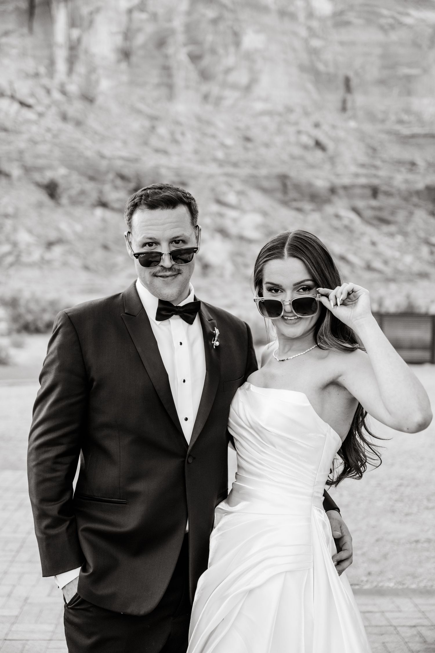 Black and white photo of a smiling bride and groom in wedding attire wearing heart-shaped sunglasses outdoors.