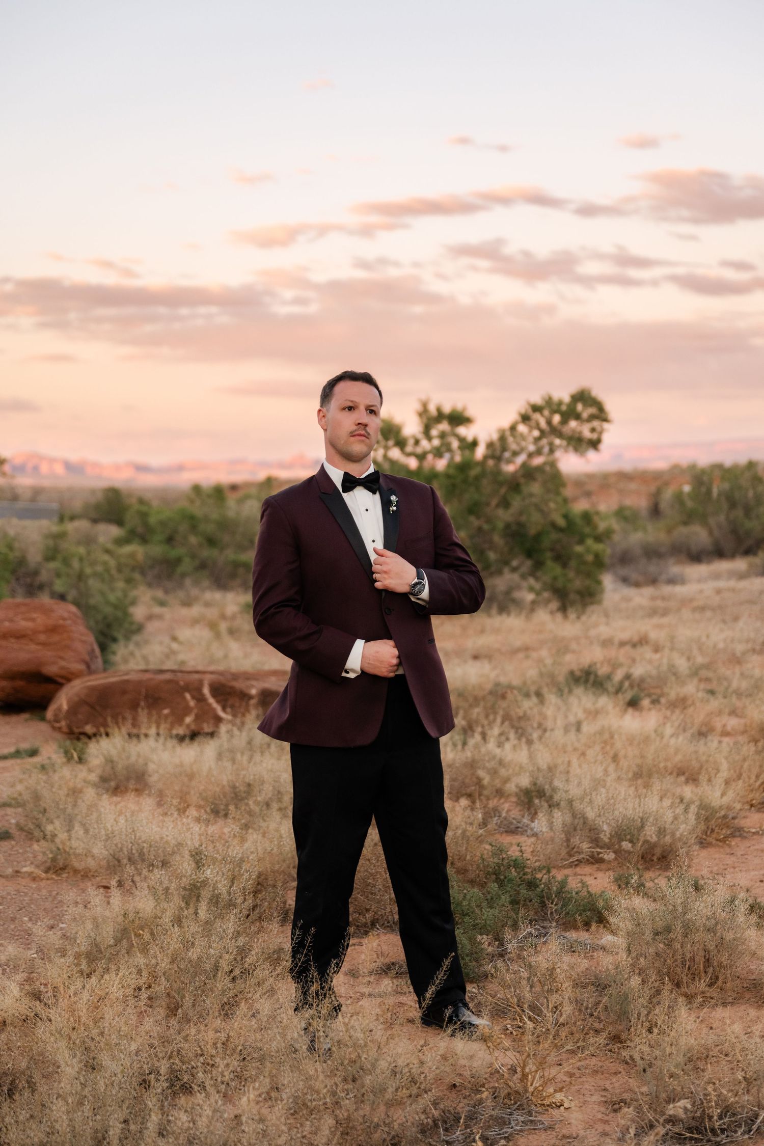 Groom in burgundy tuxedo jacket poses in desert landscape at sunset with pink sky.