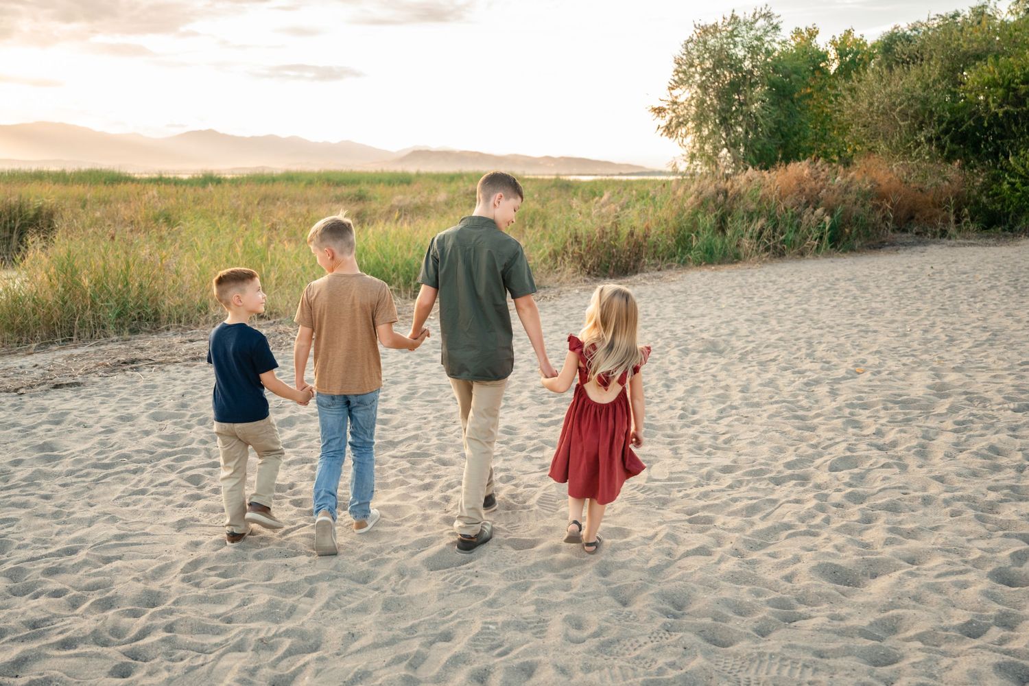 A group walks together along a sandy beach at sunset with tall grass and trees in the background.