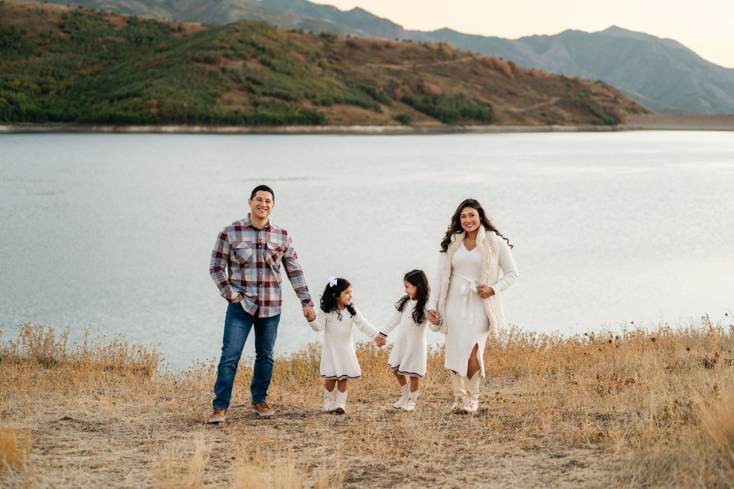 Family enjoying a playful moment together on a golden hillside with mountains and lake in the background.
