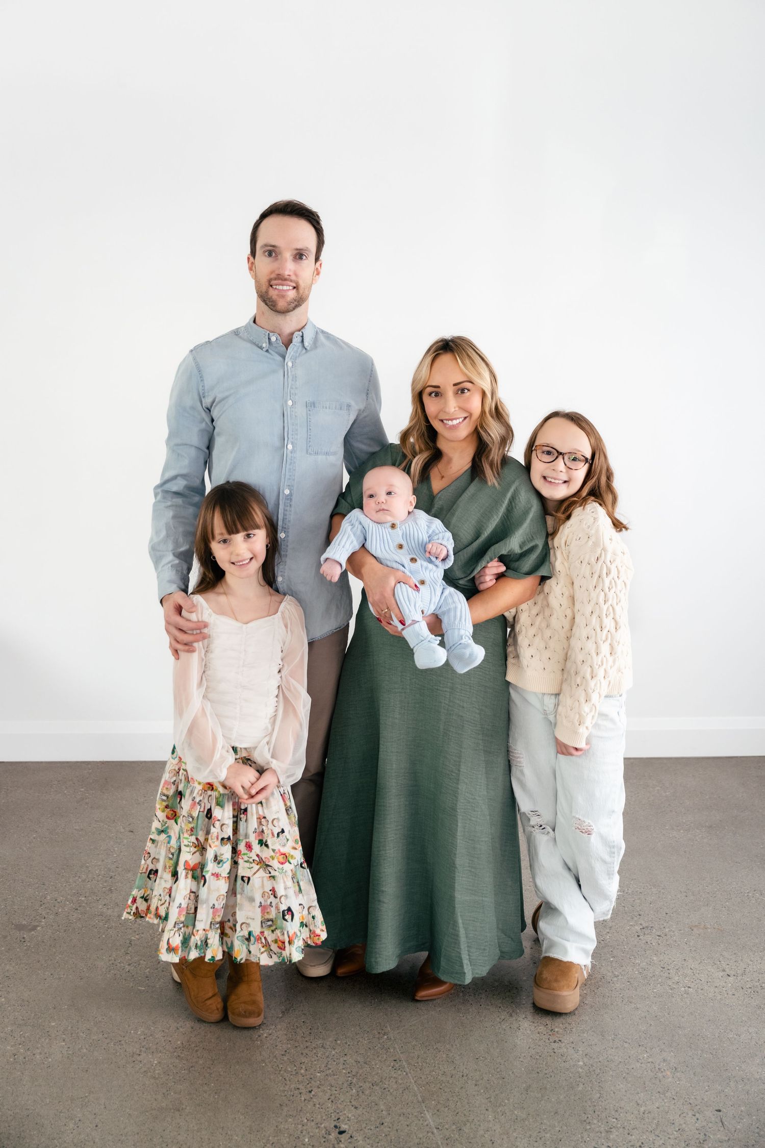 A family in coordinated casual outfits poses together against a light gray studio backdrop for a portrait session.