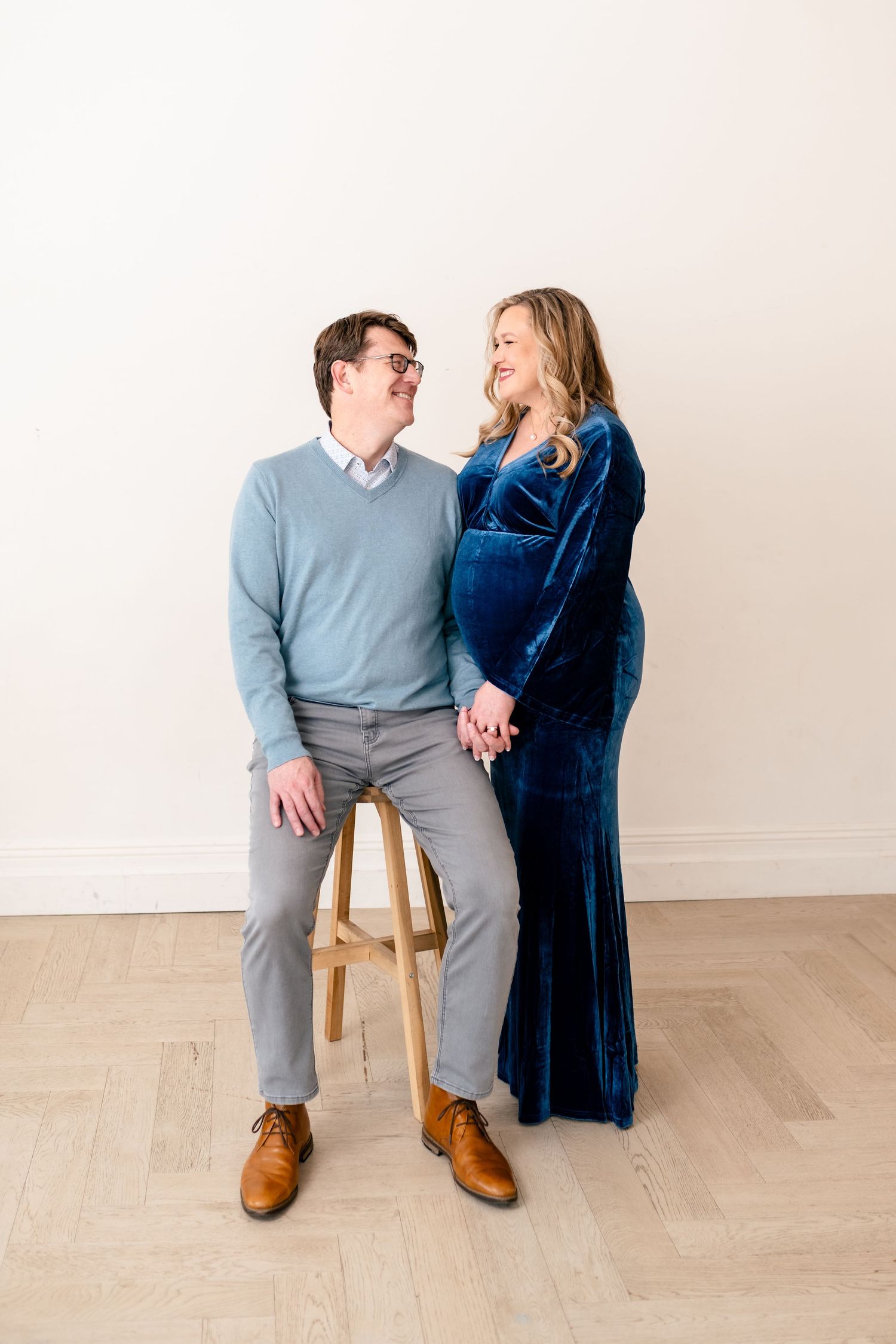 A couple in coordinating blue and gray outfits sit together on wooden stools against a neutral backdrop.