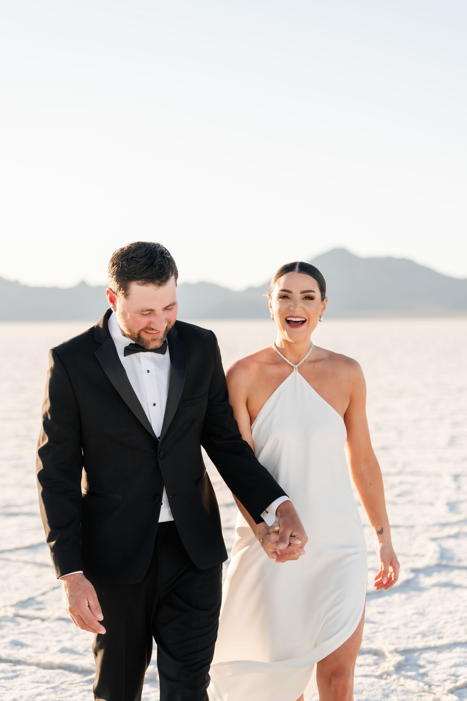 Salt Flats Photographer. A couple shares a joyful moment while walking on salt flats, with one in black tuxedo and one in white dress.