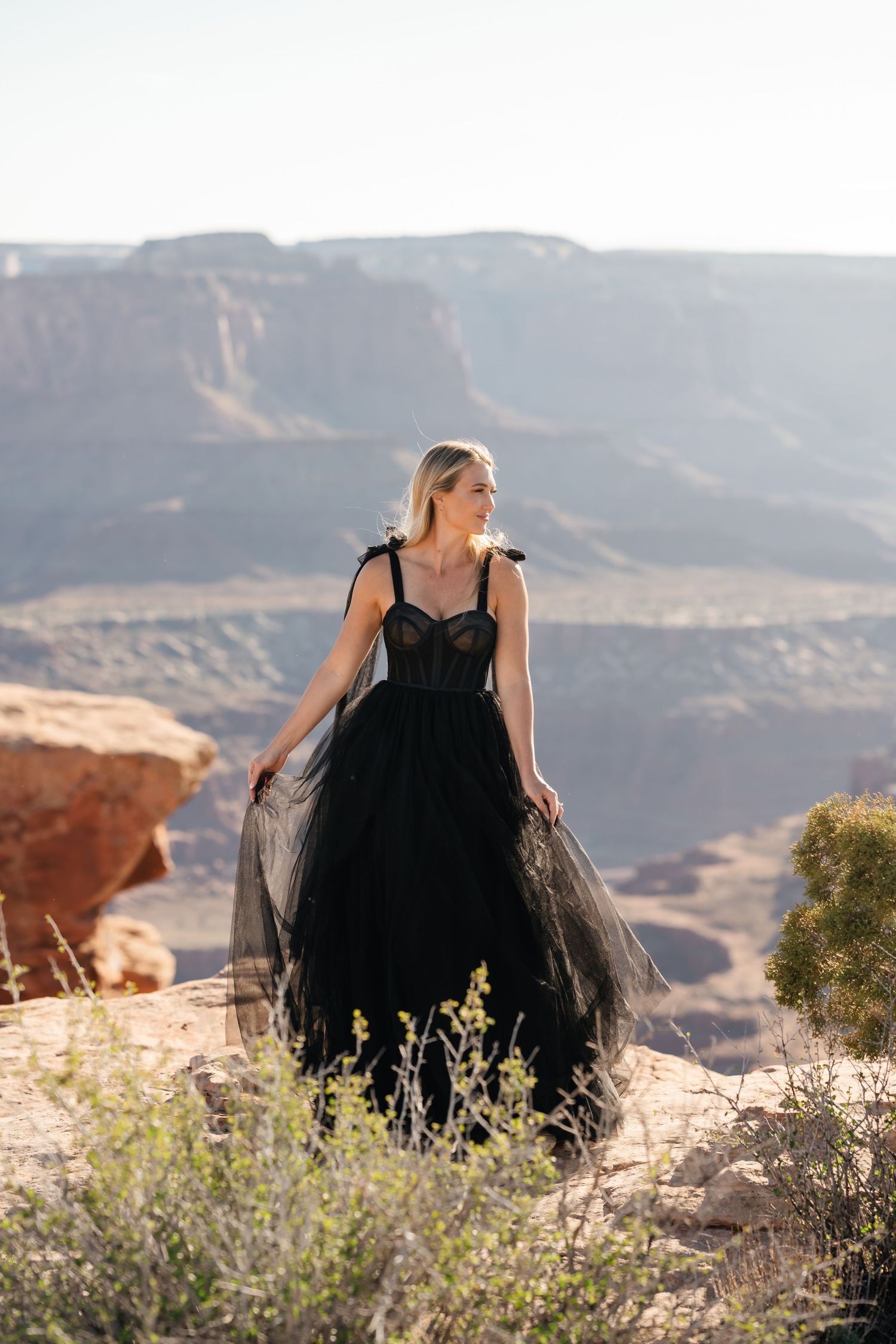 Person in a black dress sits on rocky terrain overlooking a scenic desert canyon landscape with distant mountains.