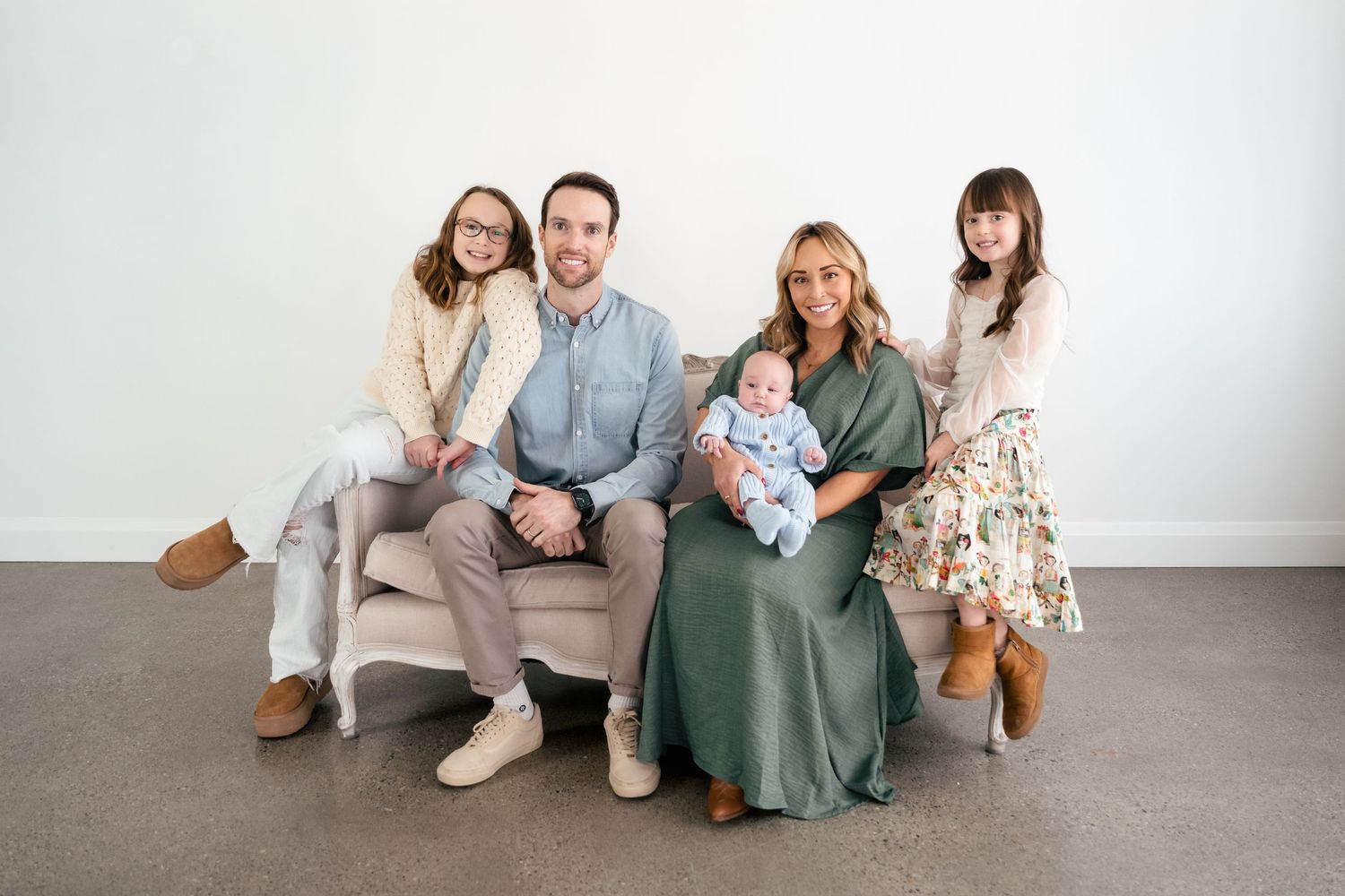 A family group sits together on a gray couch wearing coordinated neutral and sage green outfits against a white wall.