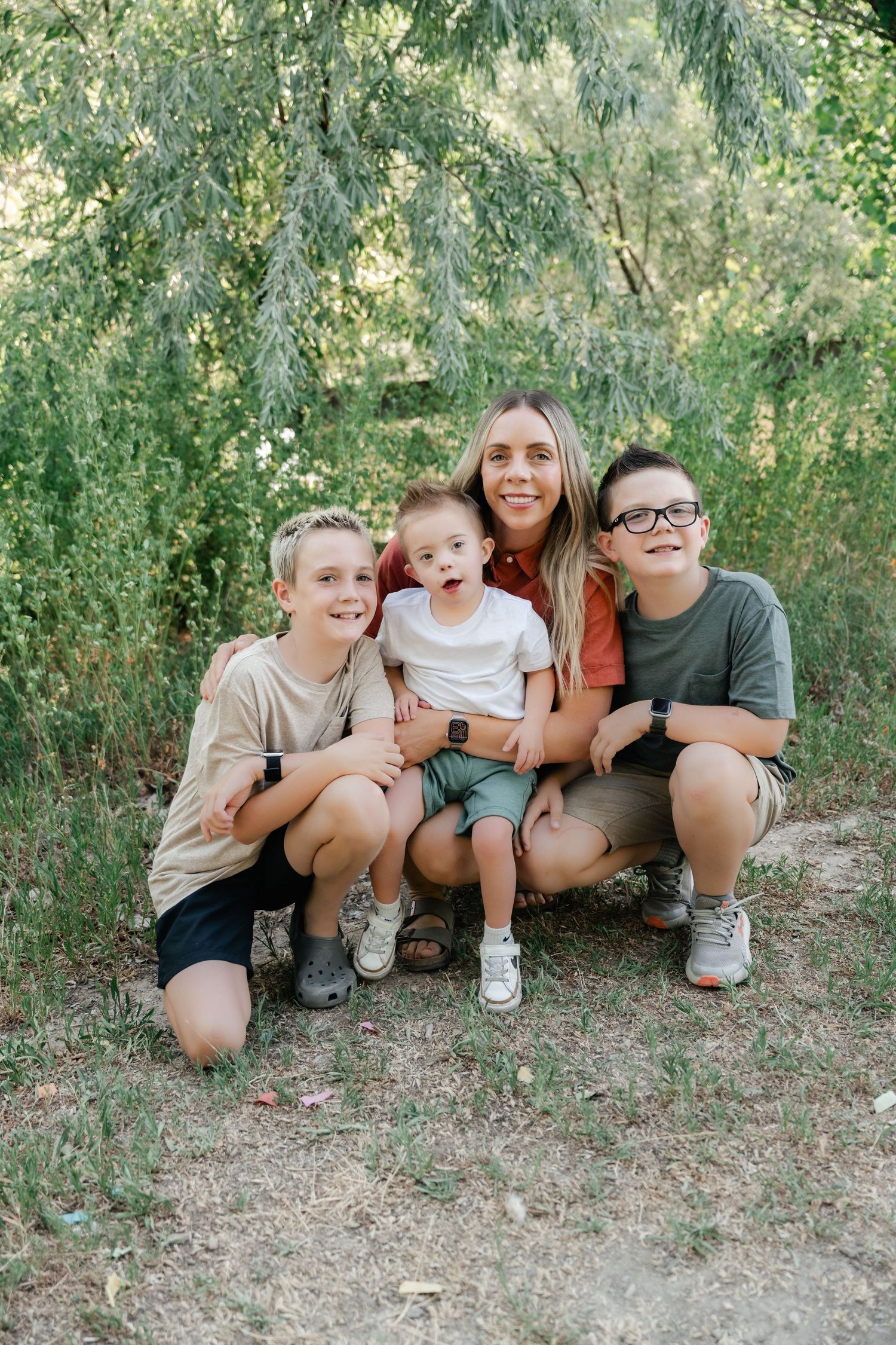 A group poses together outdoors under green trees on a sunny day in casual summer clothing.