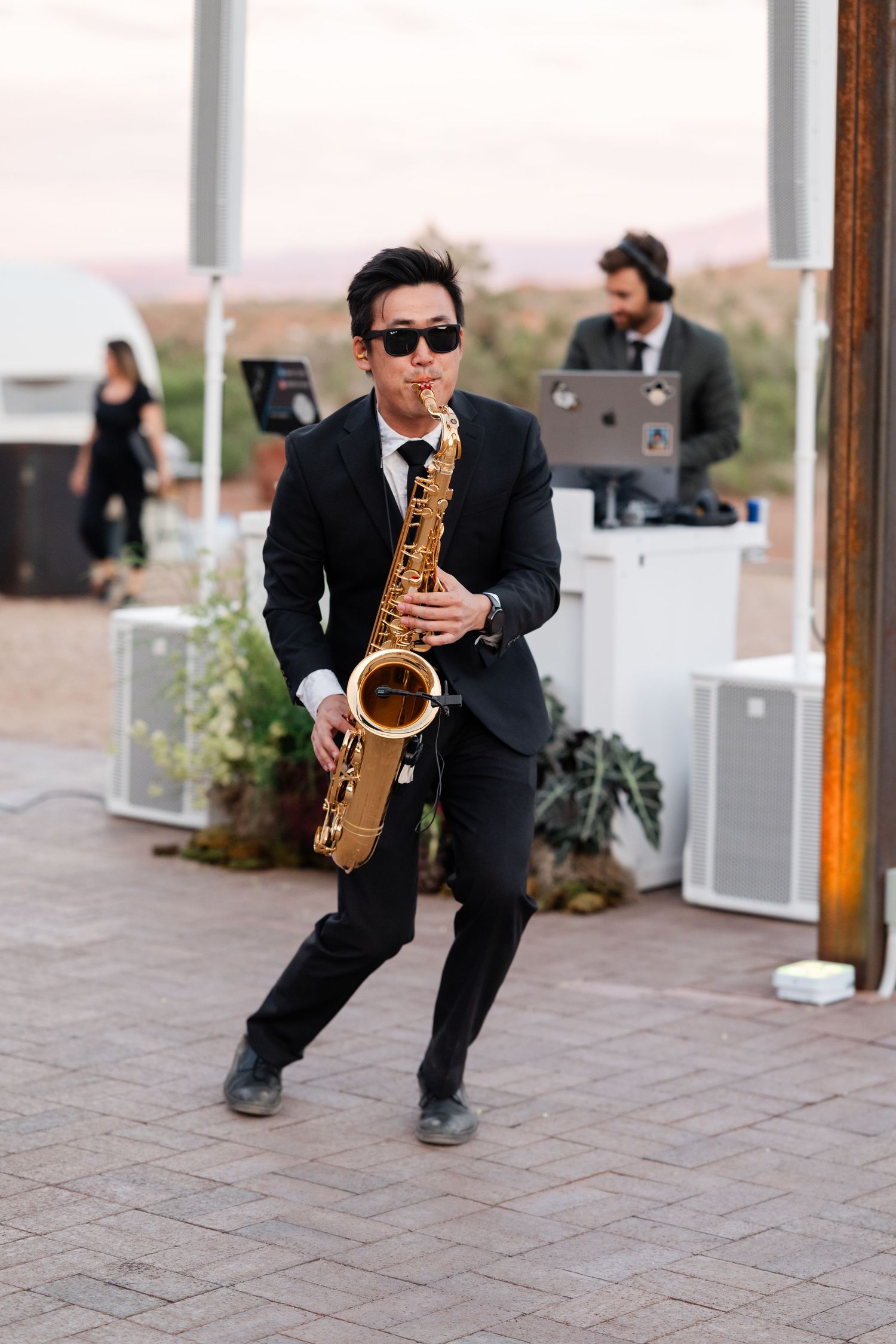 Saxophonist in black suit performs outdoors at wedding reception with DJ setup in background.