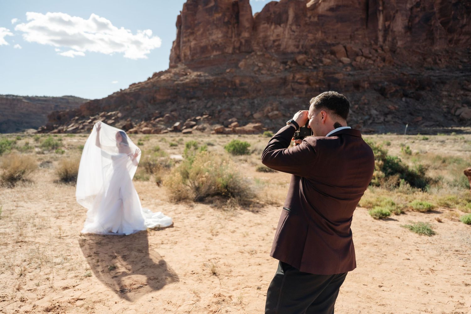 Groom photographs bride in white wedding dress and veil against red rock desert landscape in sunny outdoor elopement.
