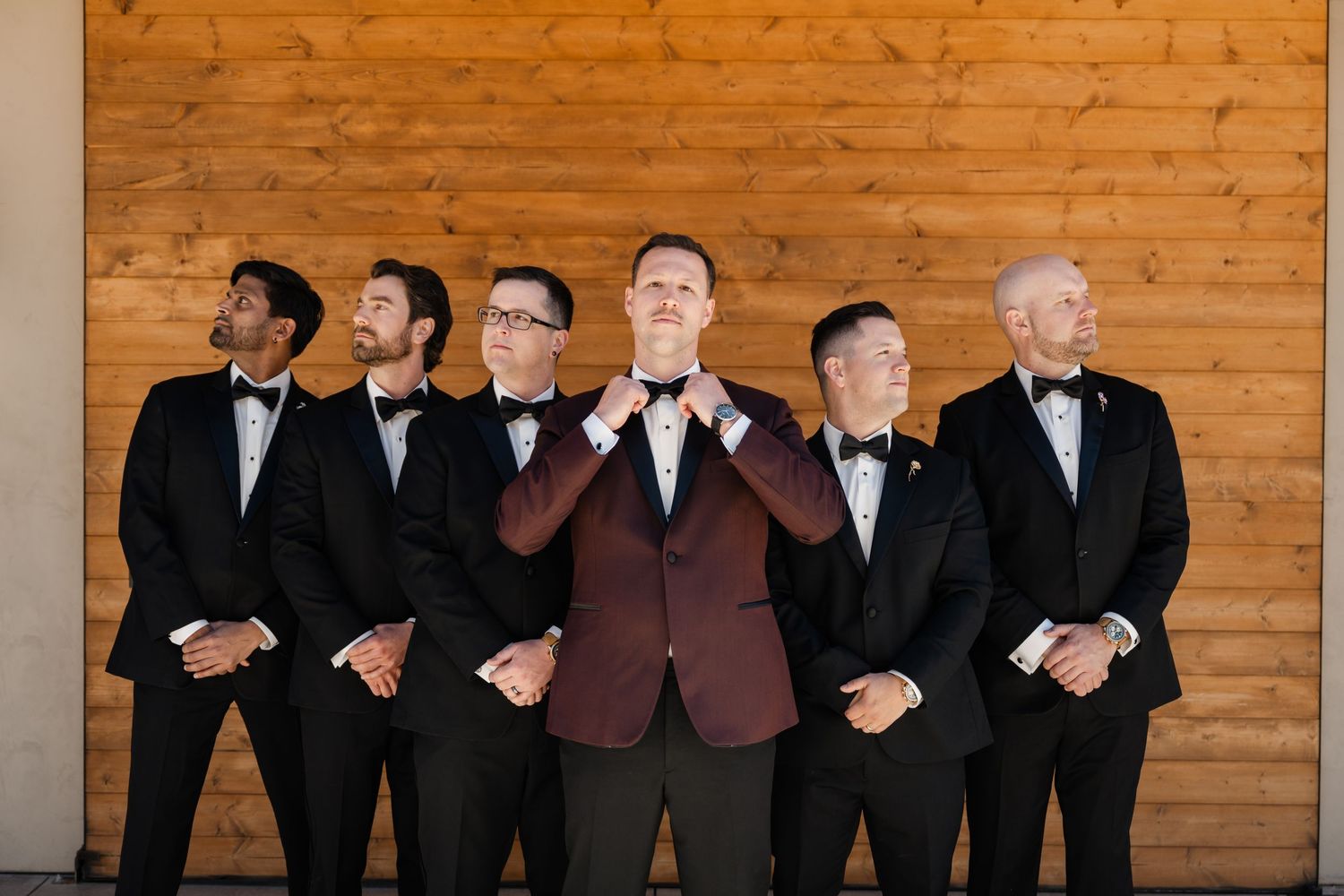 Groom in burgundy tuxedo jacket adjusting bow tie with groomsmen in black tuxedos against wooden wall.