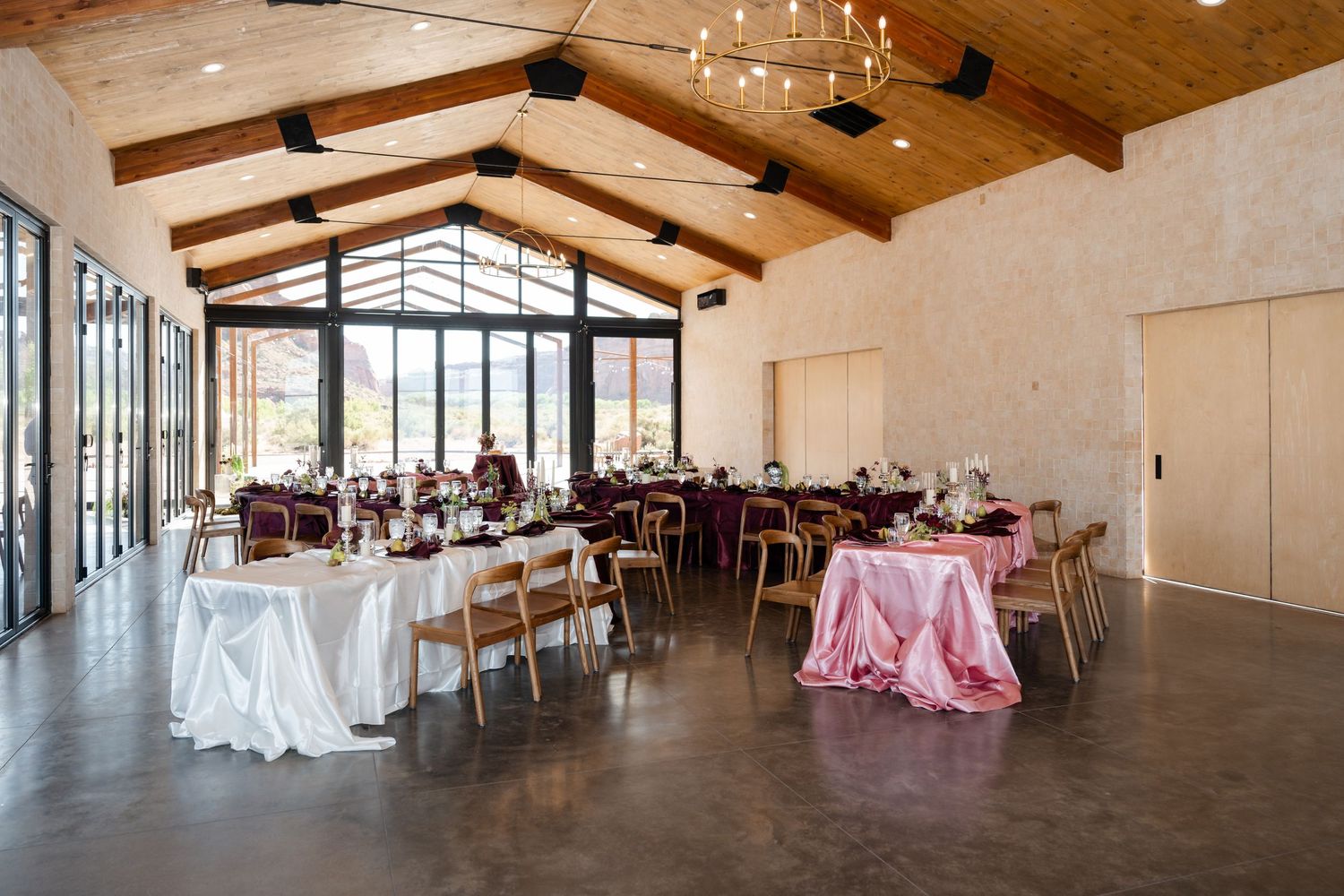 Elegant wedding reception hall with vaulted wood ceilings, pink and white draped tables, and floor-to-ceiling windows.