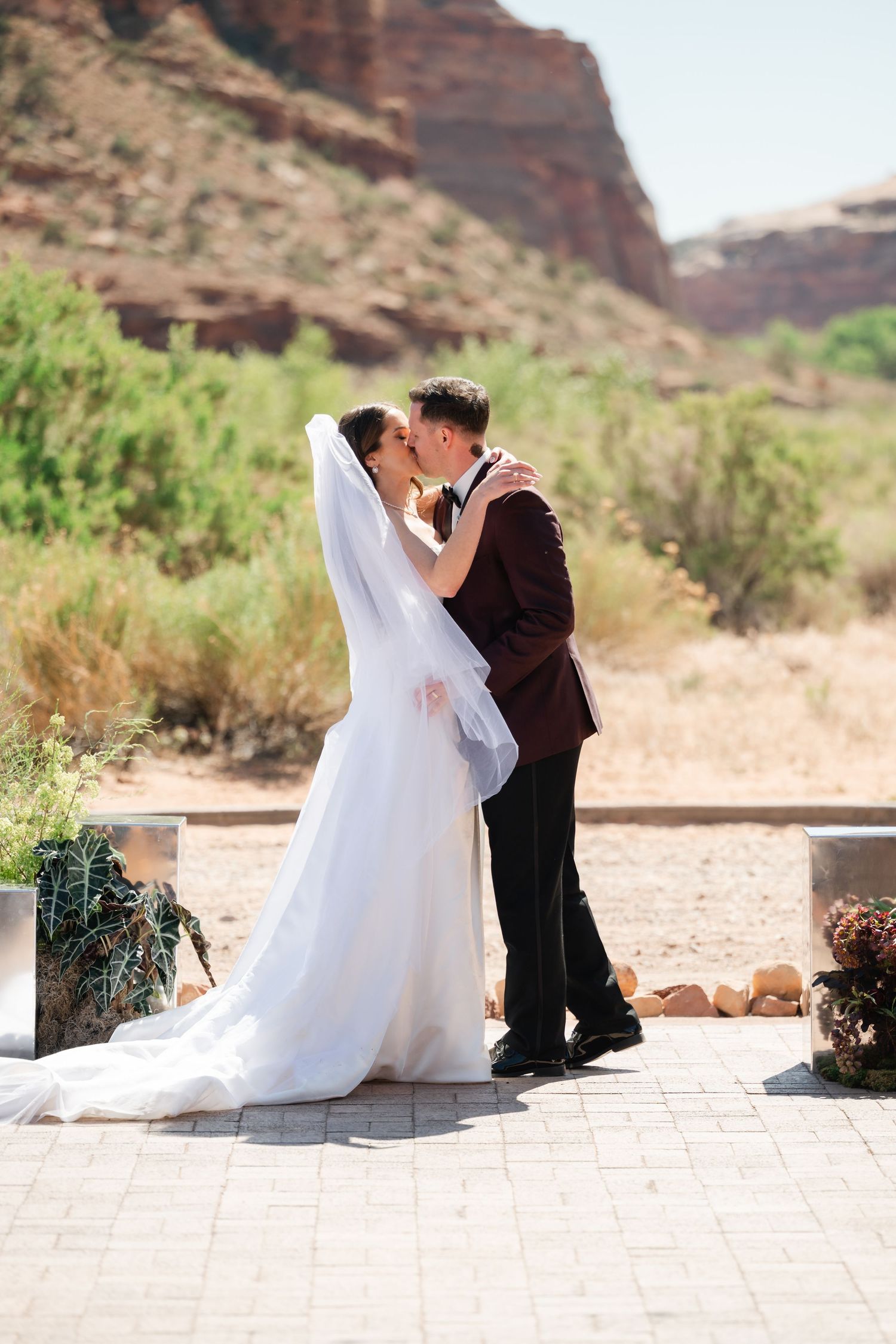 Bride and groom share a first kiss at outdoor desert wedding ceremony with red rock canyon backdrop.