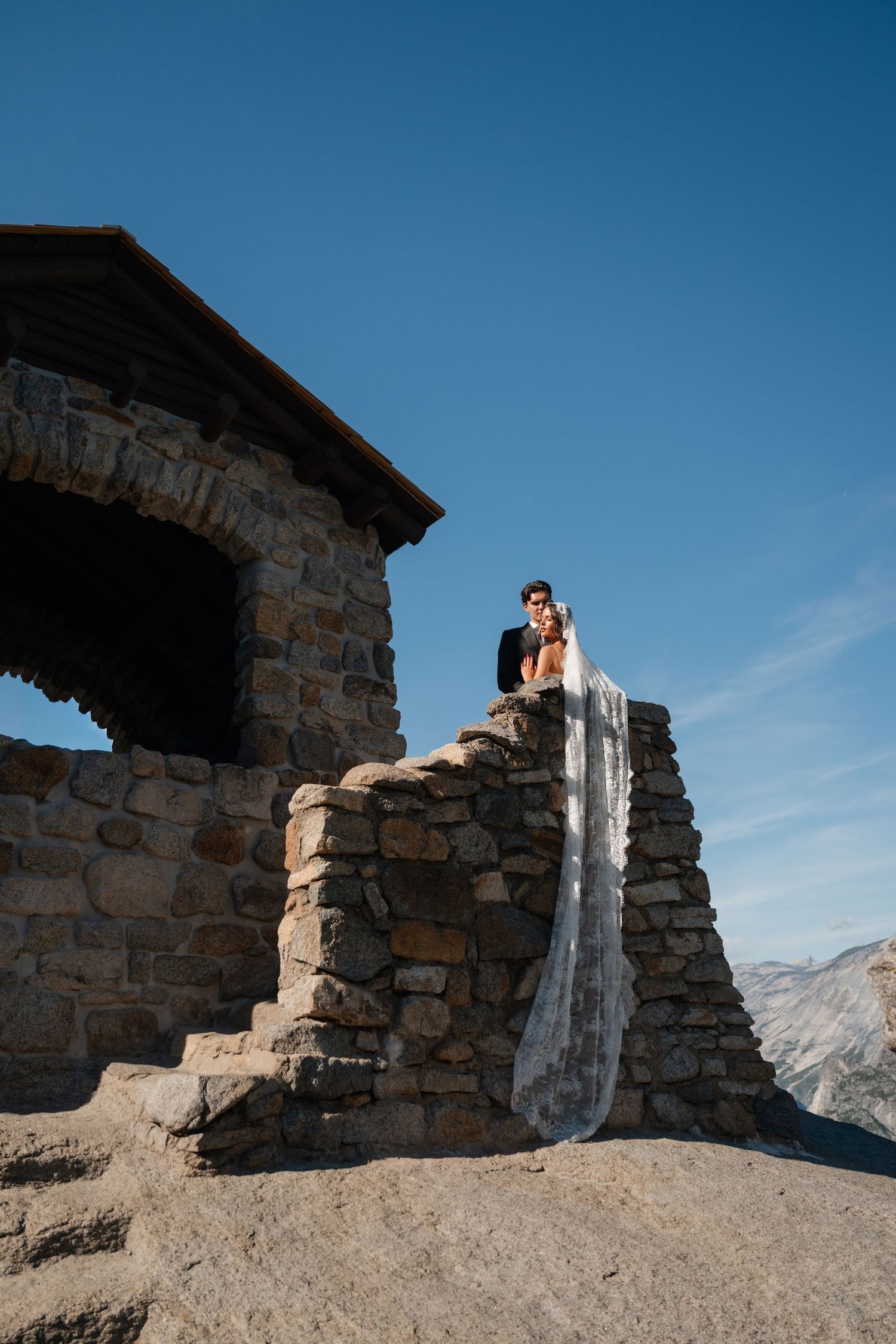 Bride and groom stand atop stone wall beside rustic mountain shelter under clear blue sky at Yosemite.
