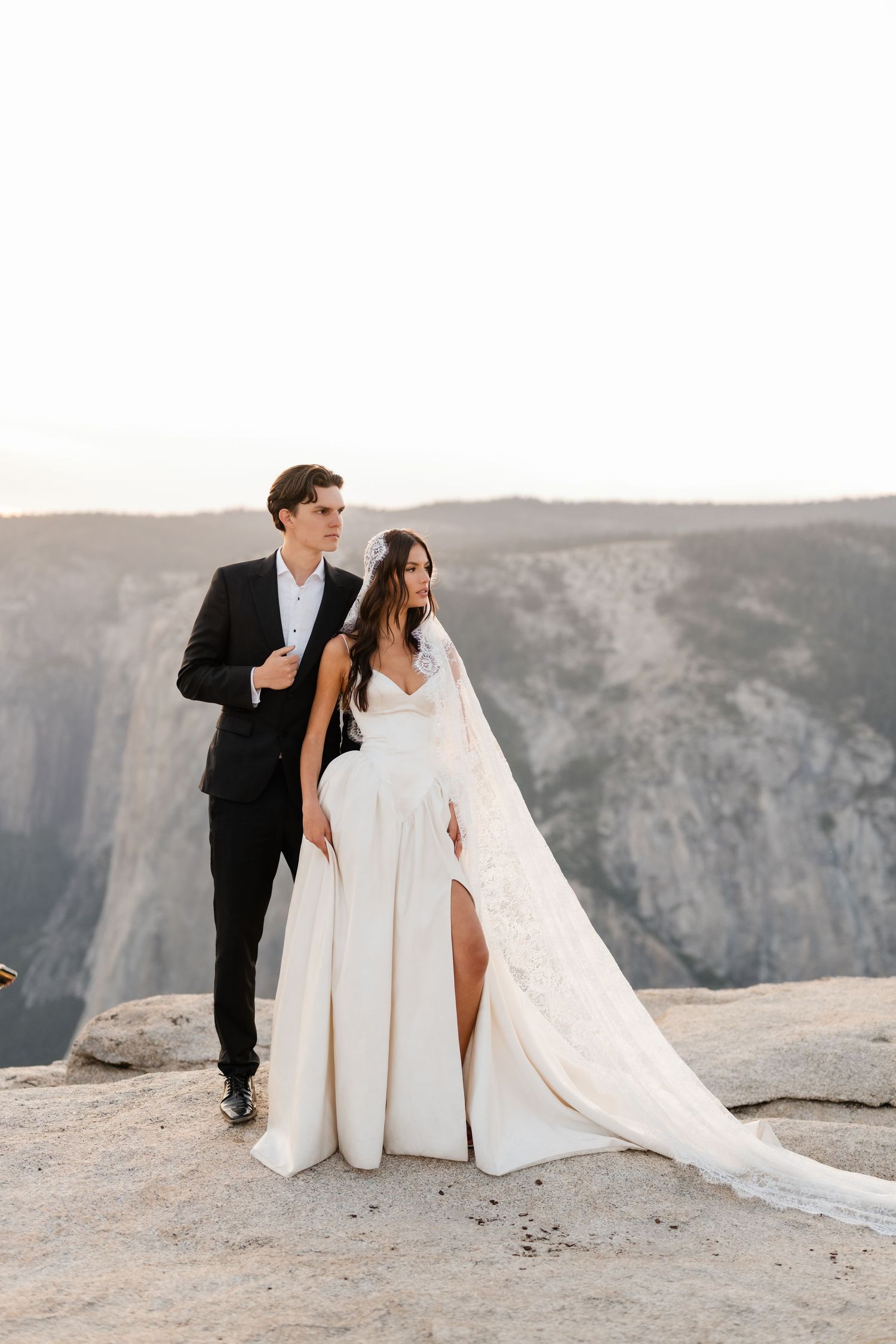 Bride and groom stand on a rocky cliff at Yosemite, her white gown and veil flowing in the breeze.